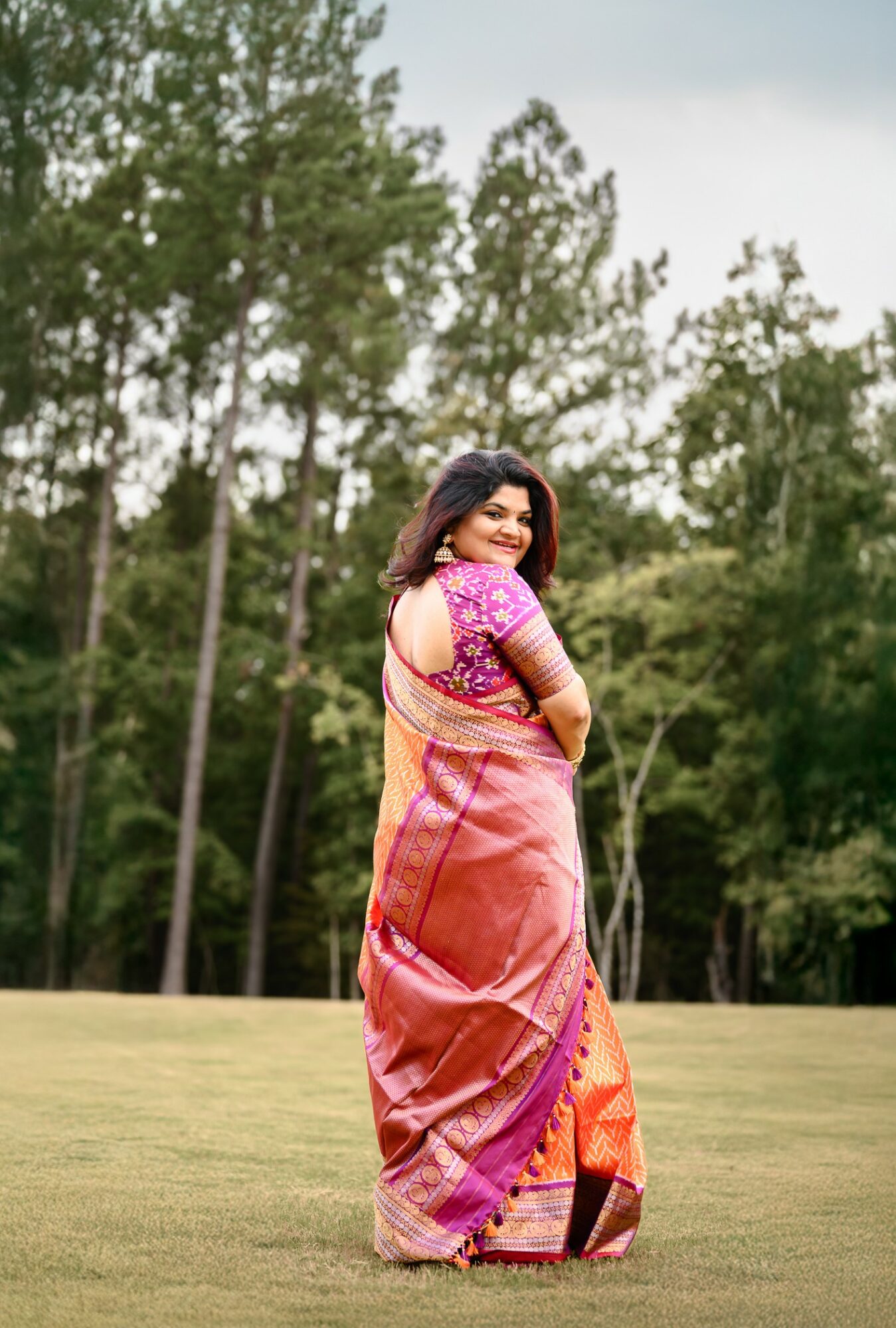 Woman in colorful saree standing outdoors on grass with trees in background, smiling, facing sideways.