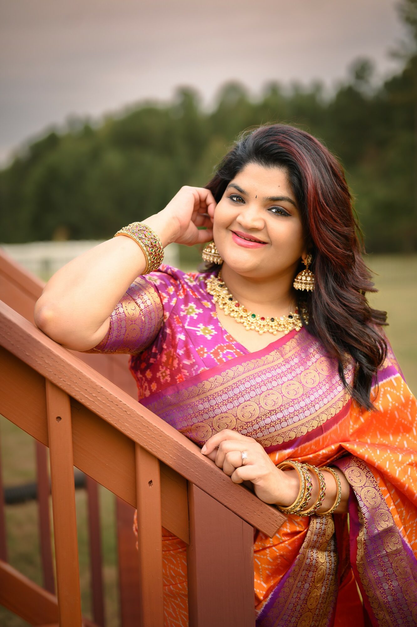 Woman in colorful traditional attire smiling outdoors, leaning on a wooden railing with trees in the background.