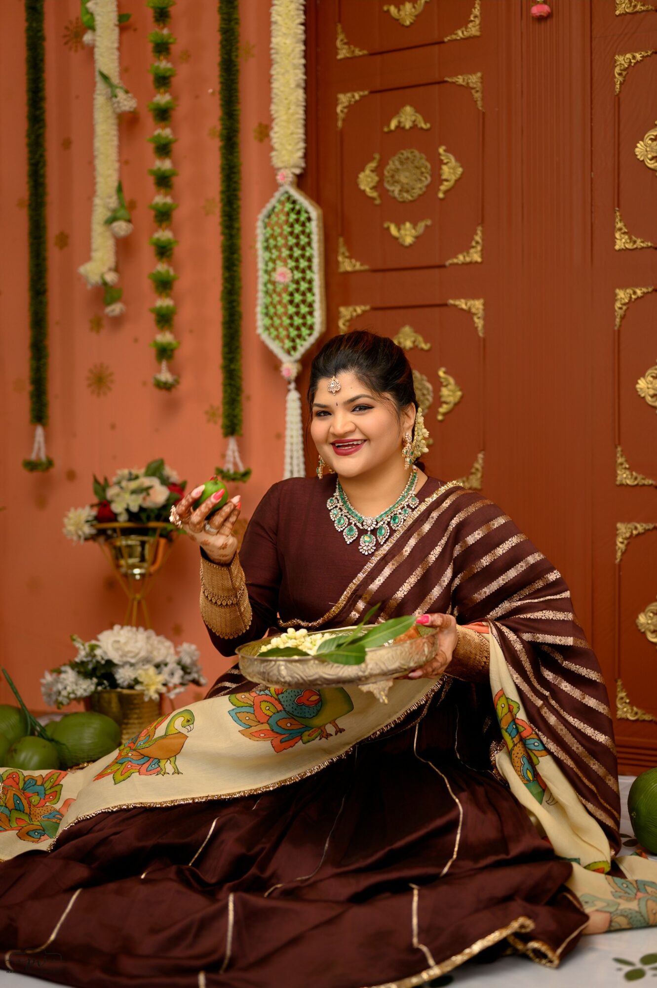 Woman in traditional attire sitting on floor, holding a tray with food, decorated with flowers and ornate background.