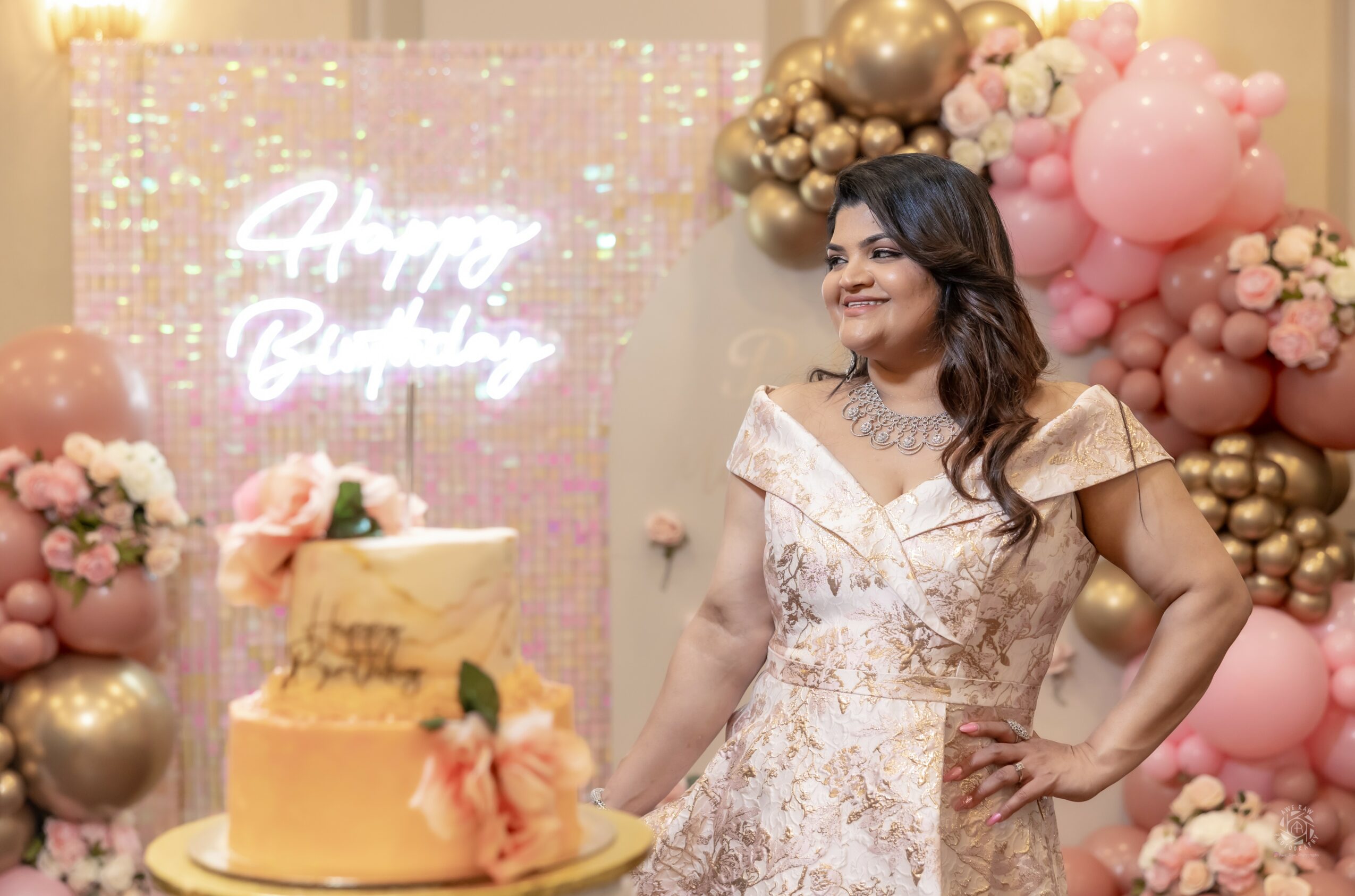 Woman in a dress standing next to a decorated cake at a celebration with pink and gold balloons and a neon sign background.
