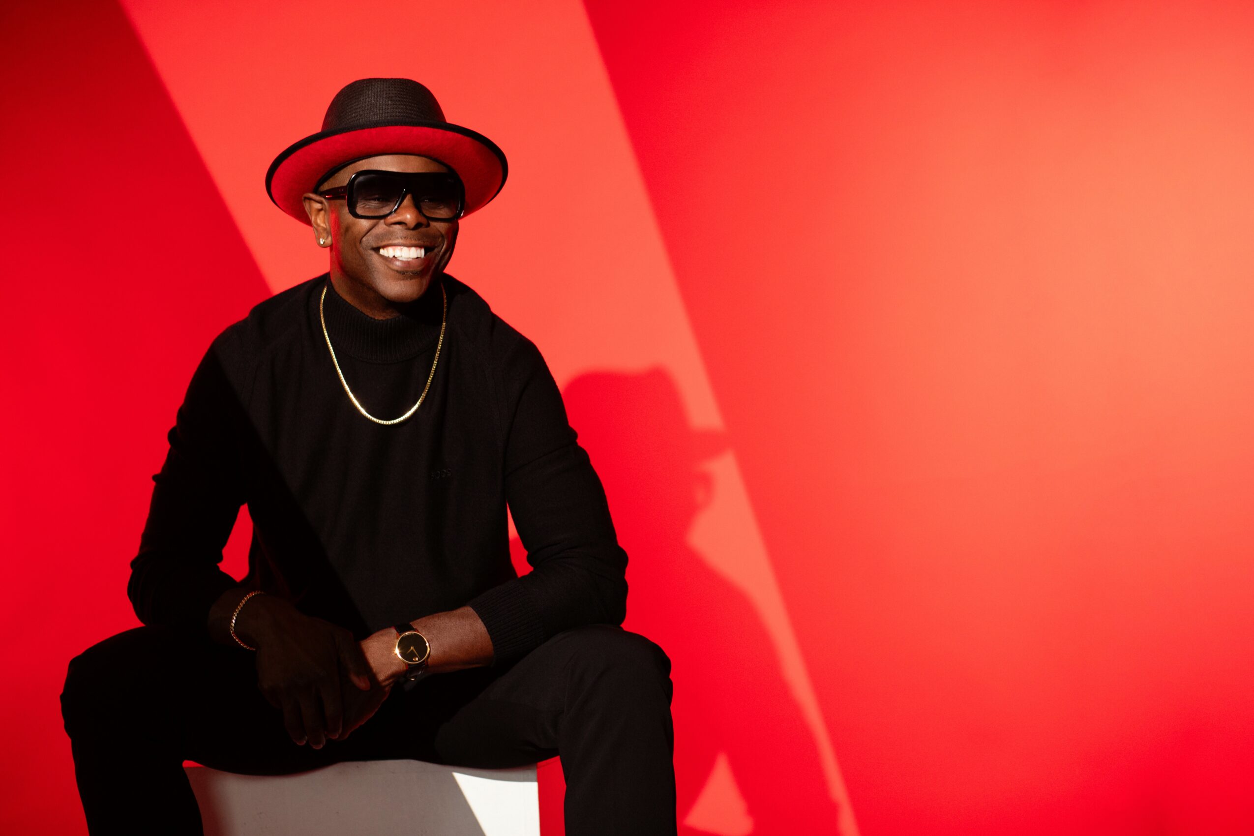 Person wearing a black outfit, red hat, and sunglasses, smiling against a red background.