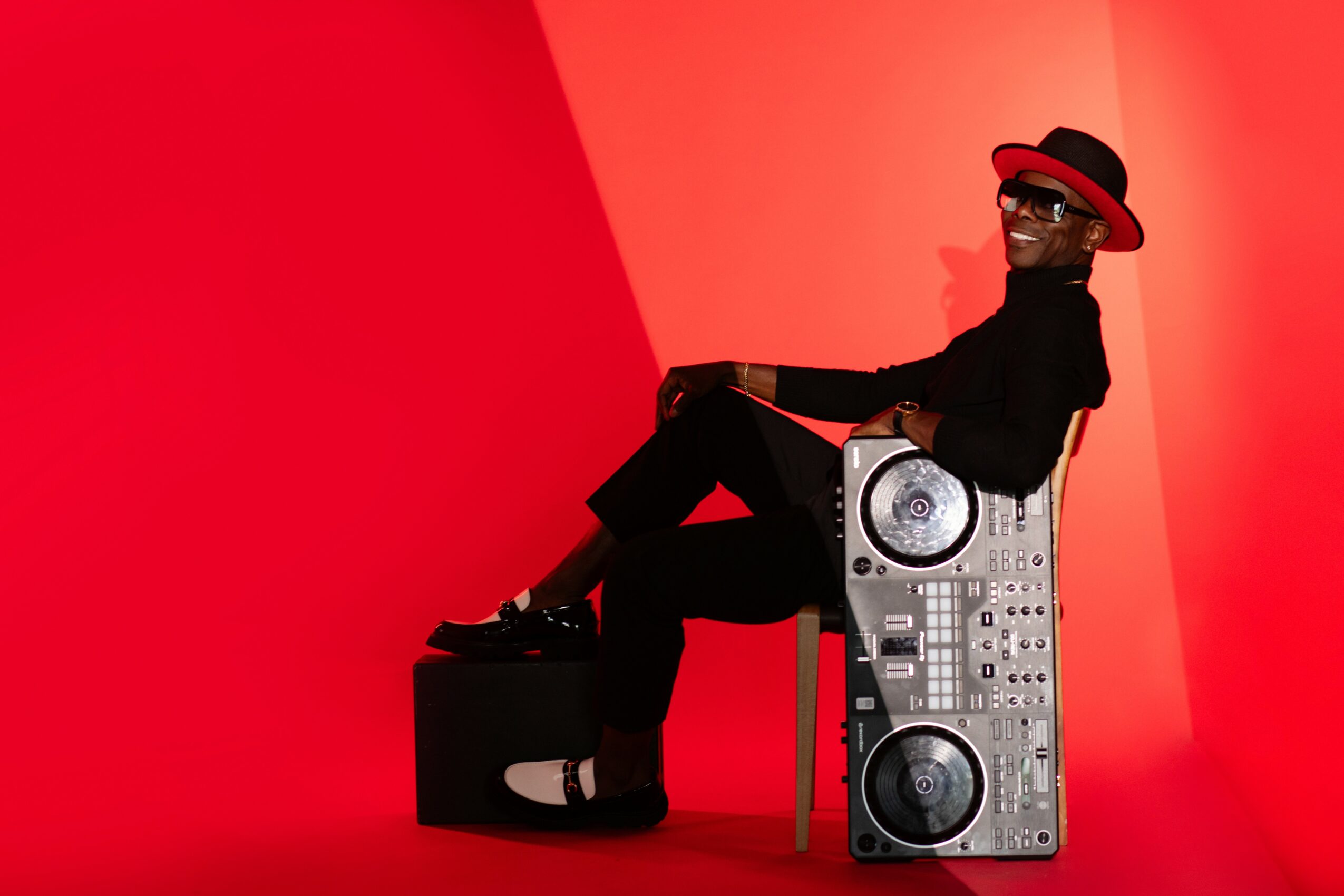 Person wearing a hat and sunglasses sitting on a speaker with a red background.