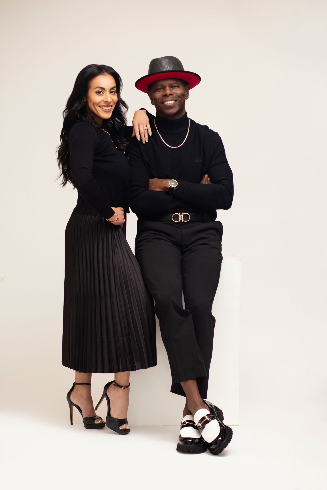 Two women stand together, smiling, one with long dark hair and the other wearing a hat, against a plain background.