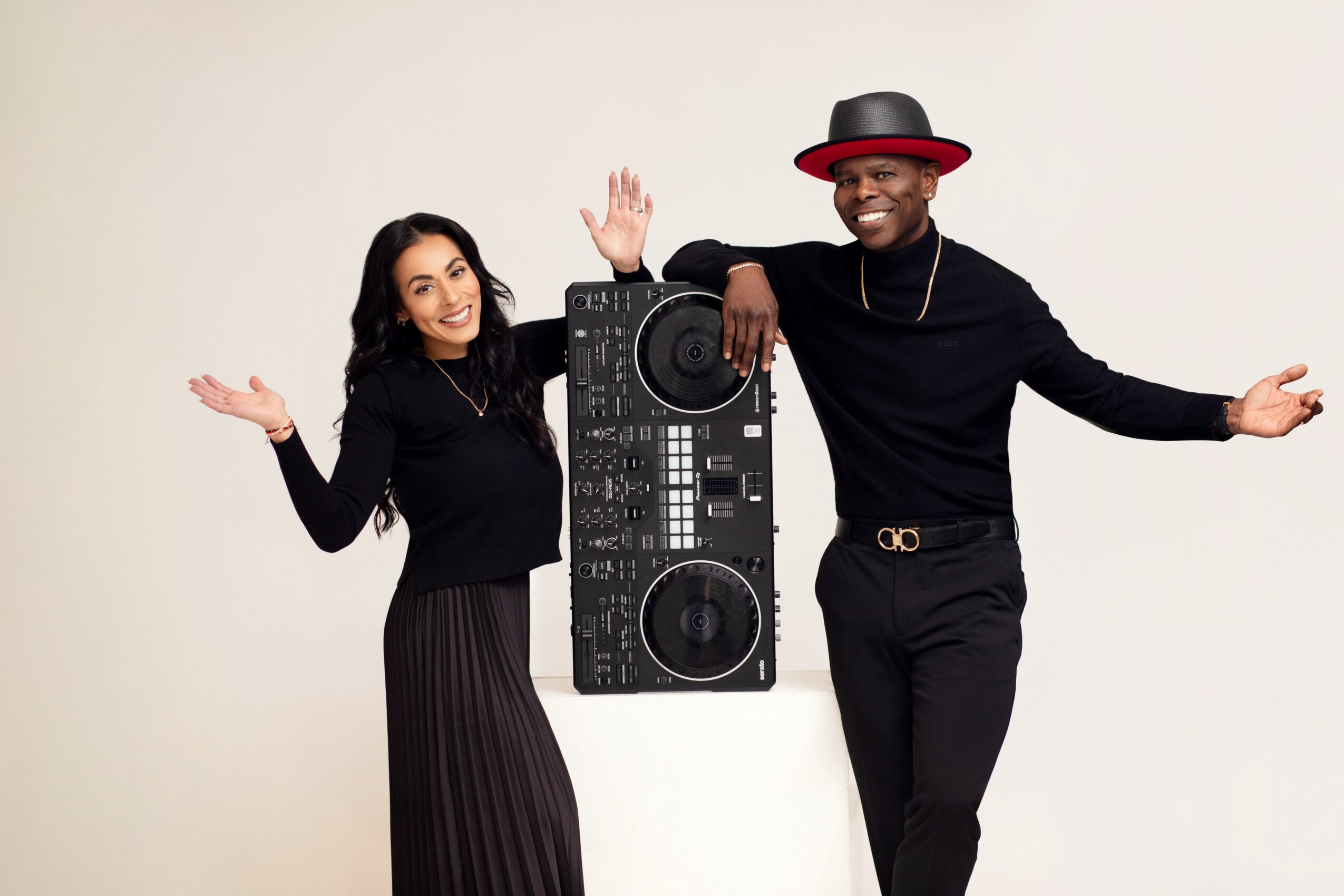 Two people stand with a DJ controller between them, smiling and waving, against a plain background.