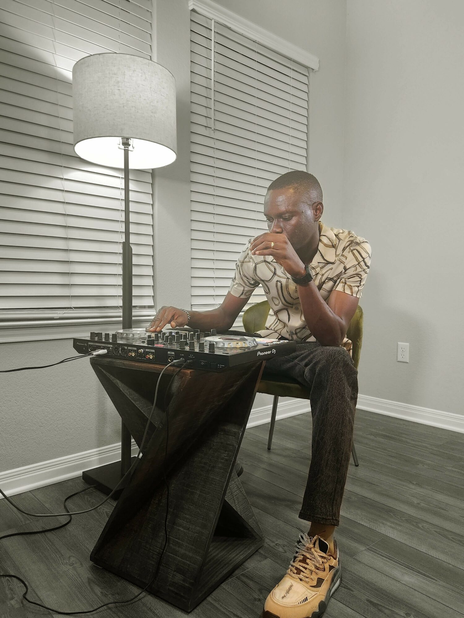 Person with glasses and patterned shirt using DJ equipment in a room with blinds and a floor lamp.