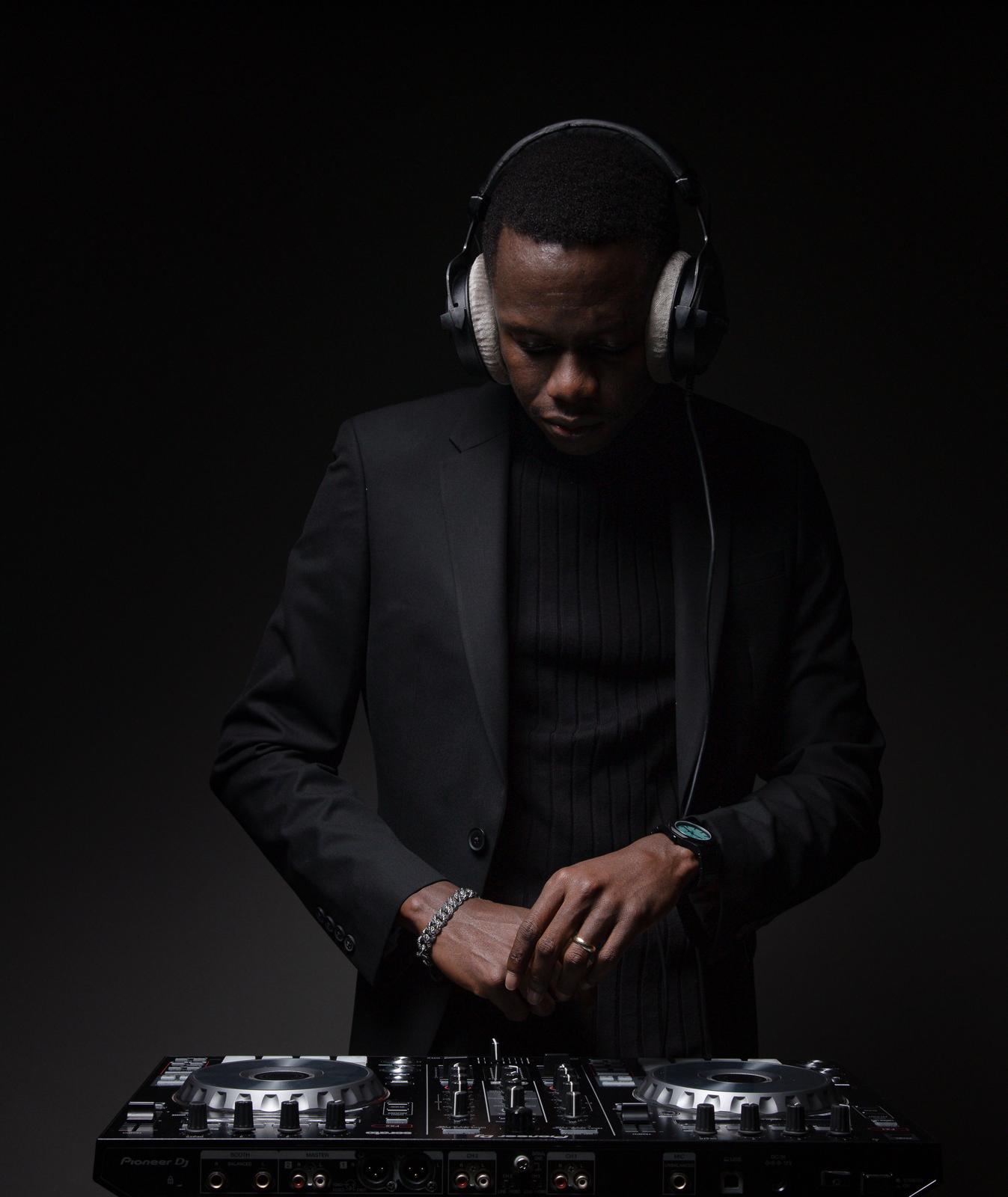 Person wearing headphones and a black suit, standing behind DJ equipment, looking down, dark background.