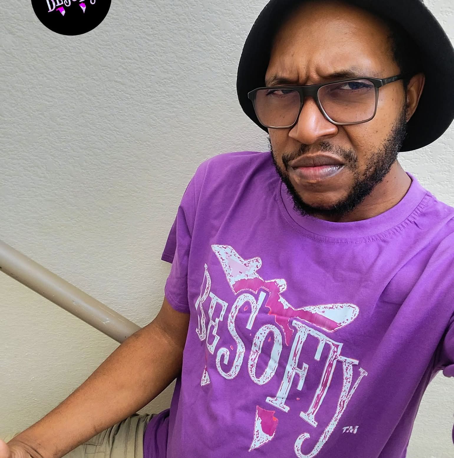 Man wearing glasses, black hat, and purple T-shirt with white text, standing outdoors against a light gray wall.