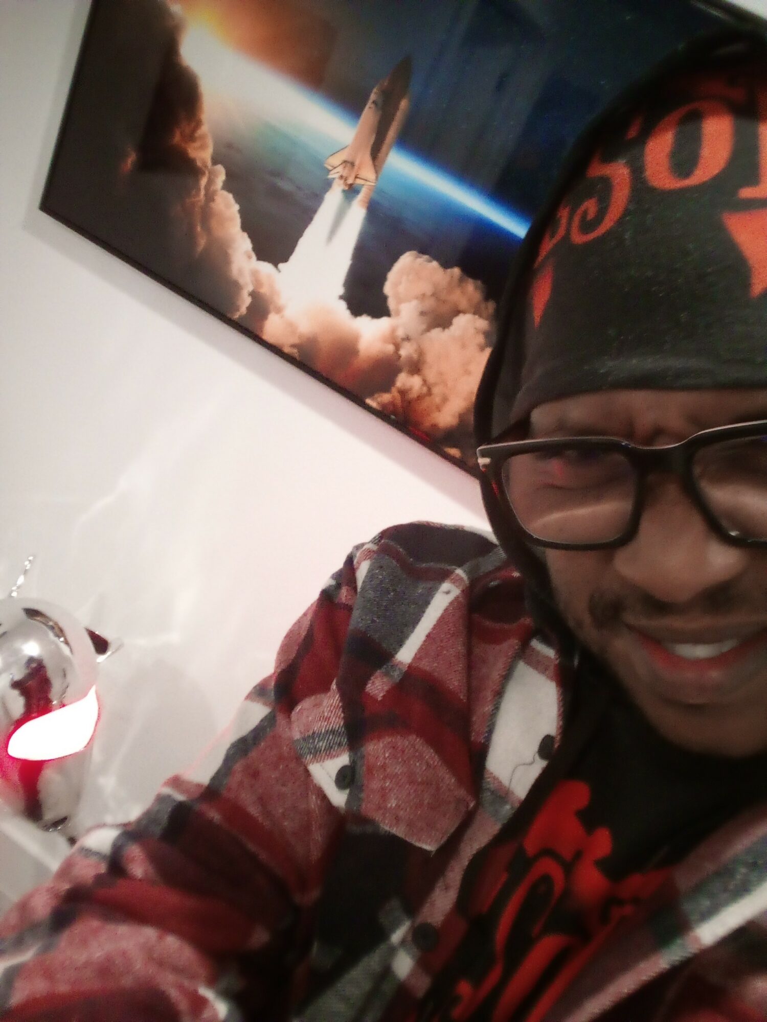 Person wearing glasses, a beanie, and a plaid shirt smiling in front of a space-themed poster with a rocket launch.