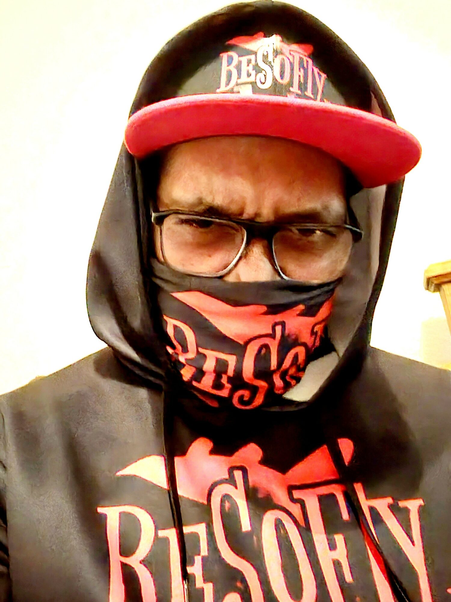 Person wearing glasses, a black hoodie, a red and black face mask, and a red cap with white text, looking at the camera.