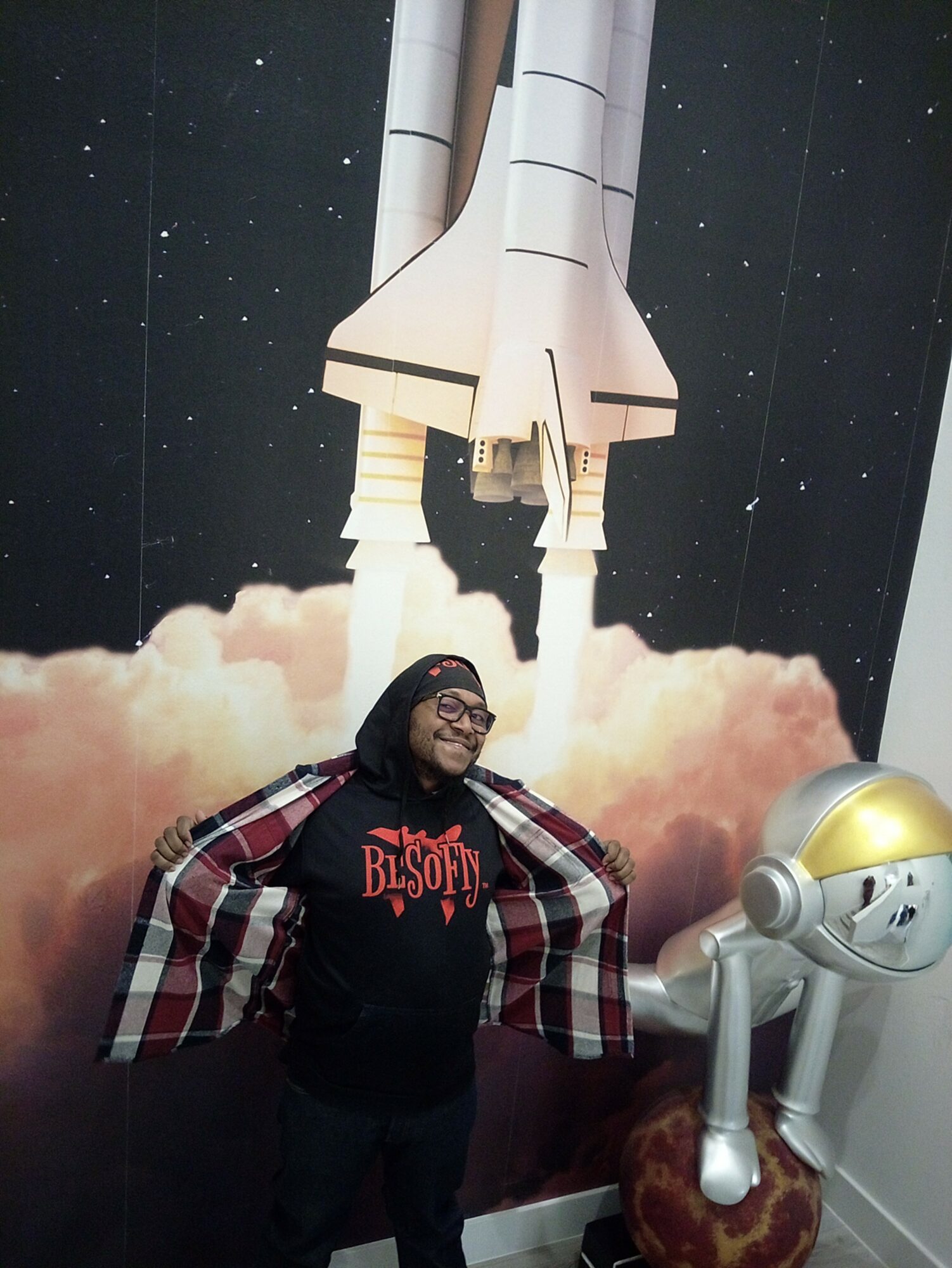 Person smiling in front of a space shuttle launch mural, with a robotic arm model nearby.