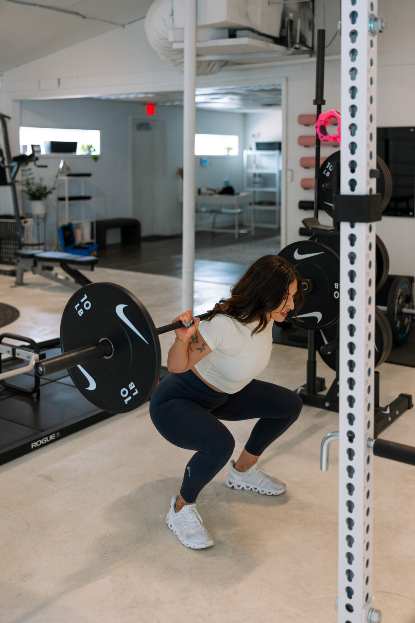 Woman performing squat exercise with barbell in gym, weight plates and equipment visible in background.