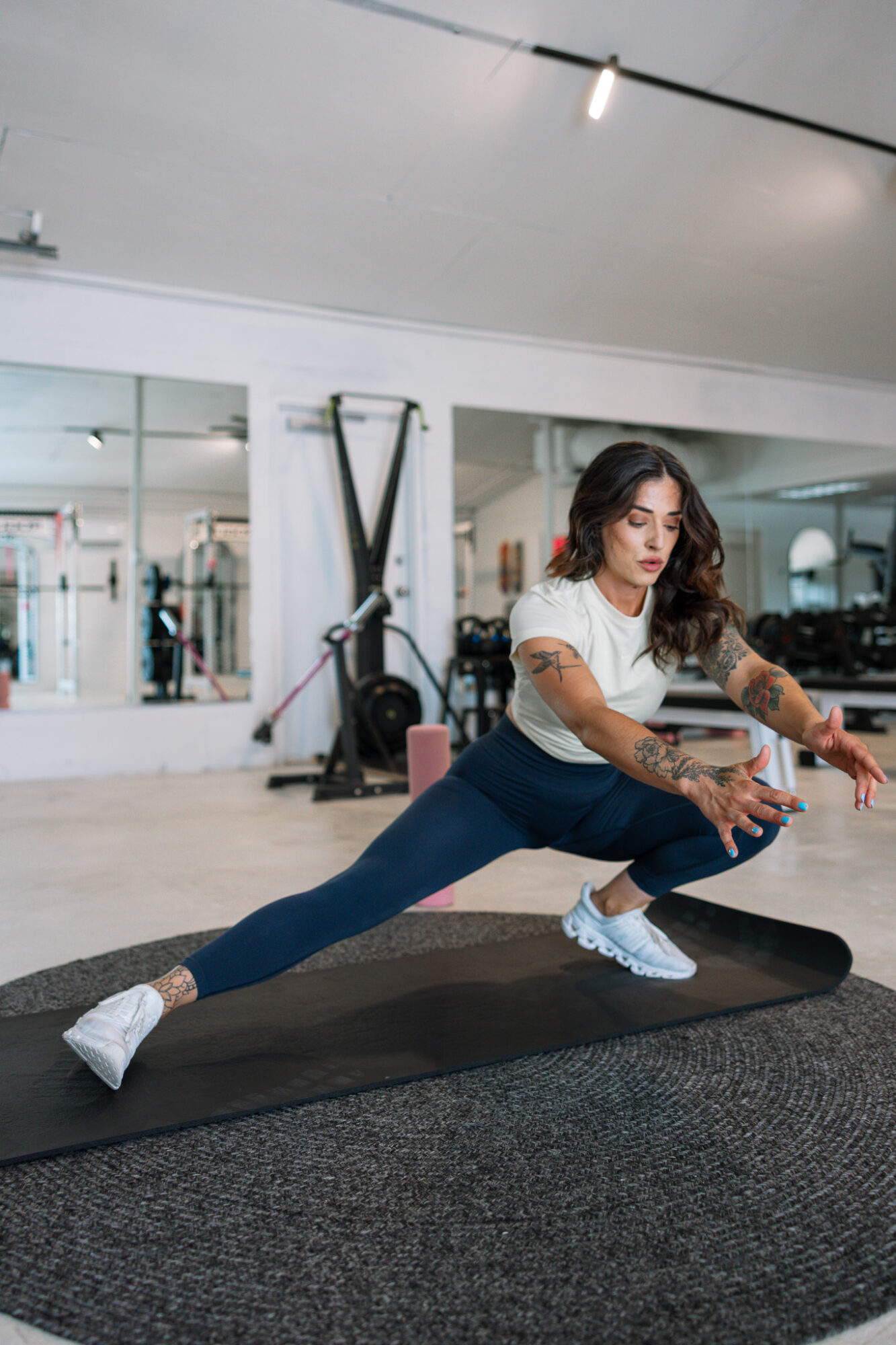 Woman stretching in gym, lunging with one leg extended and arms reaching forward, on a black mat.