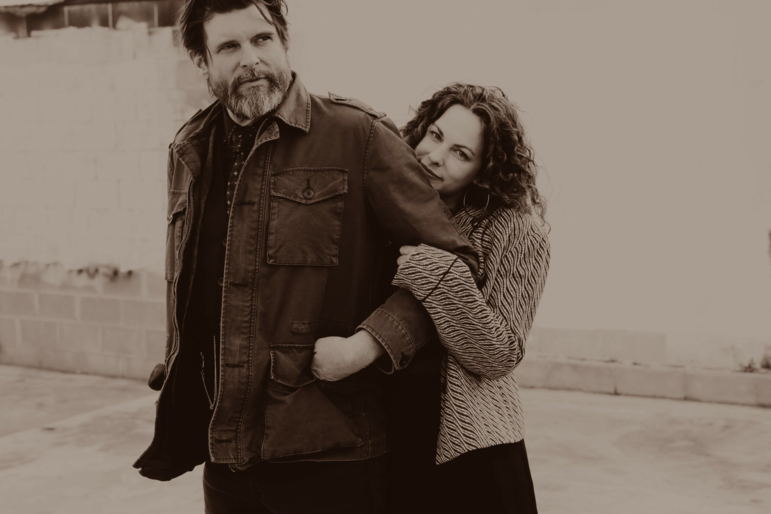 A man and woman stand outdoors, with the woman hugging the man from behind, both smiling.