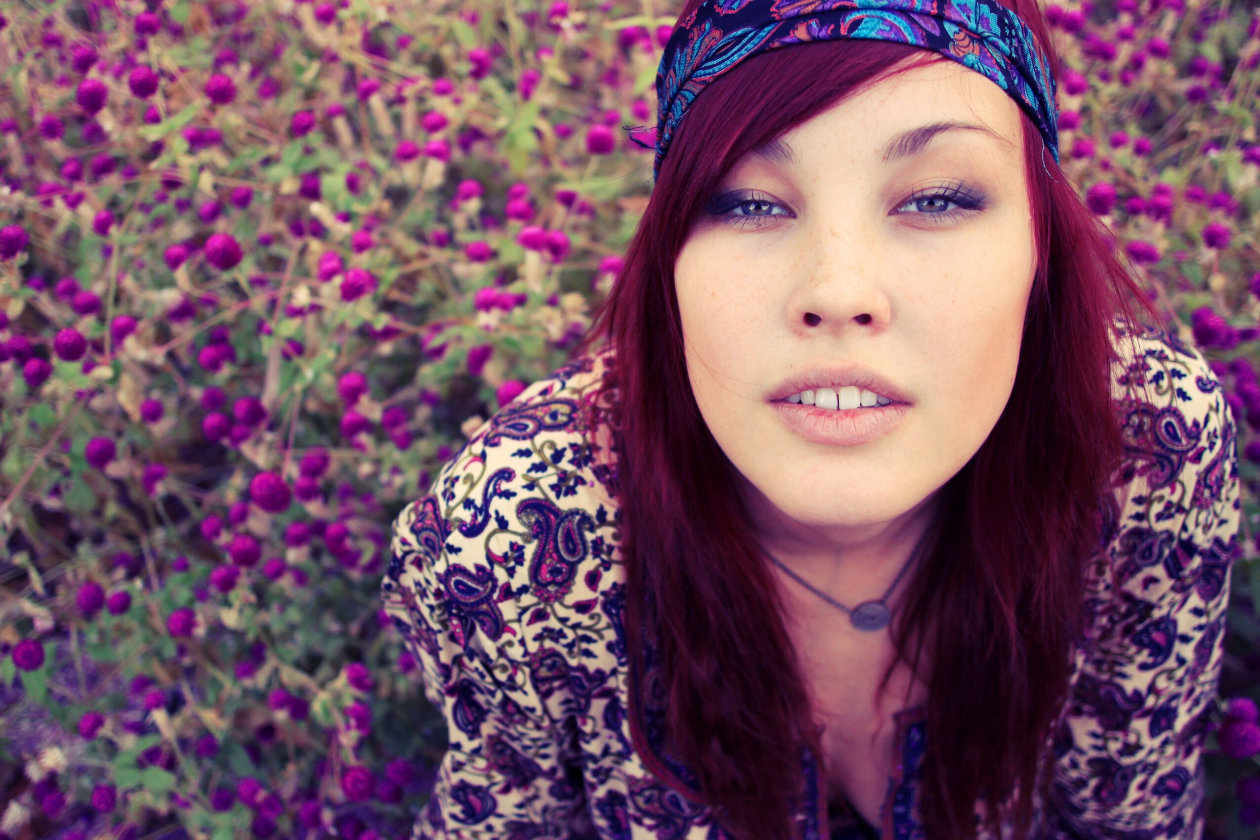 Young woman with purple hair and patterned headband outdoors among purple flowers, looking at the camera.
