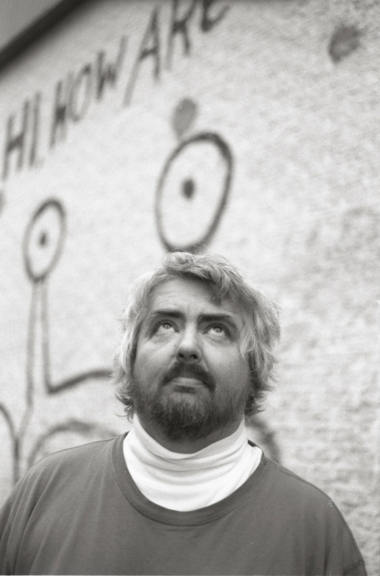 Man with beard looking upward in front of a wall with abstract drawings and text, black and white.