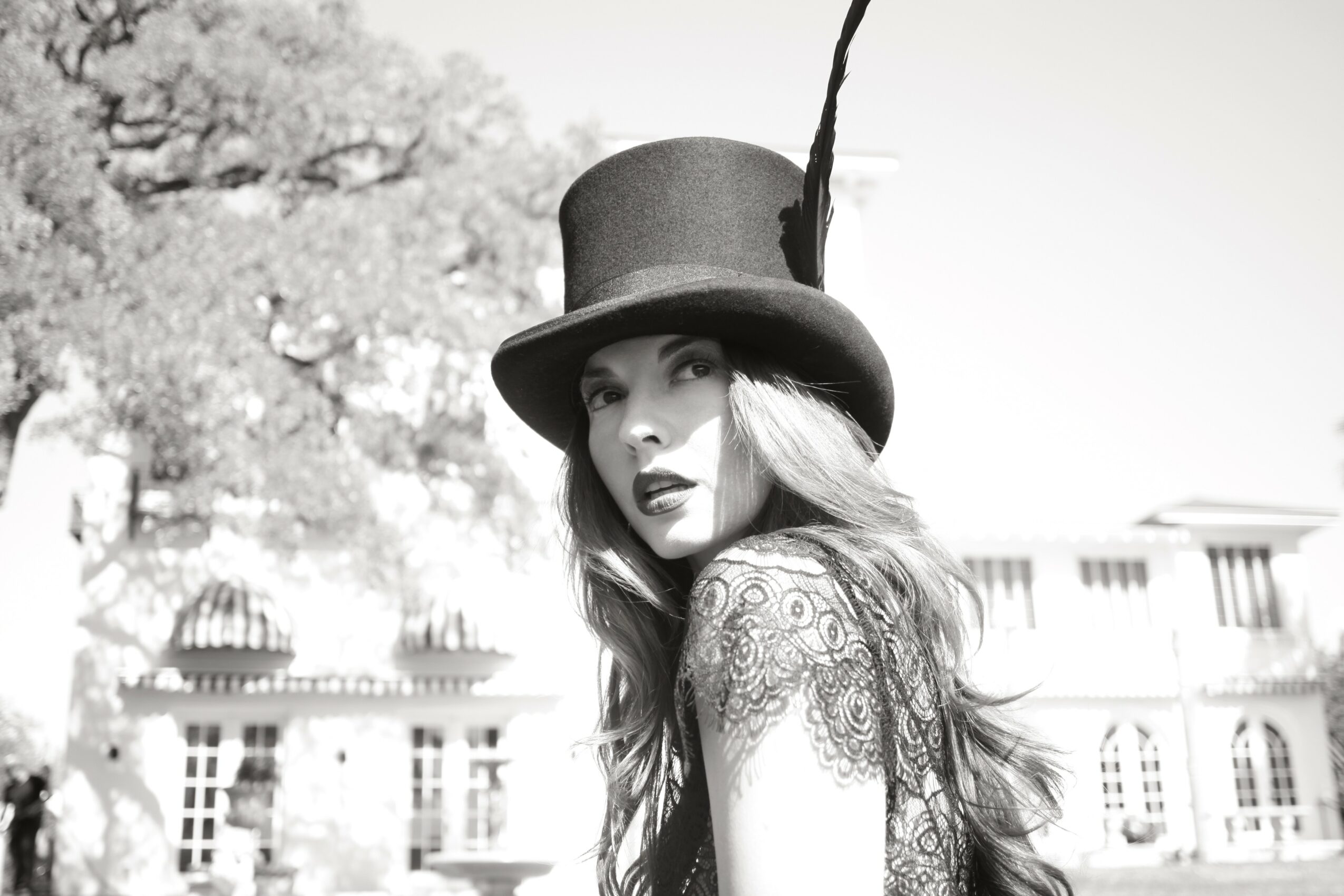 Woman wearing a top hat with a feather, looking at the camera, with buildings and trees in the background.