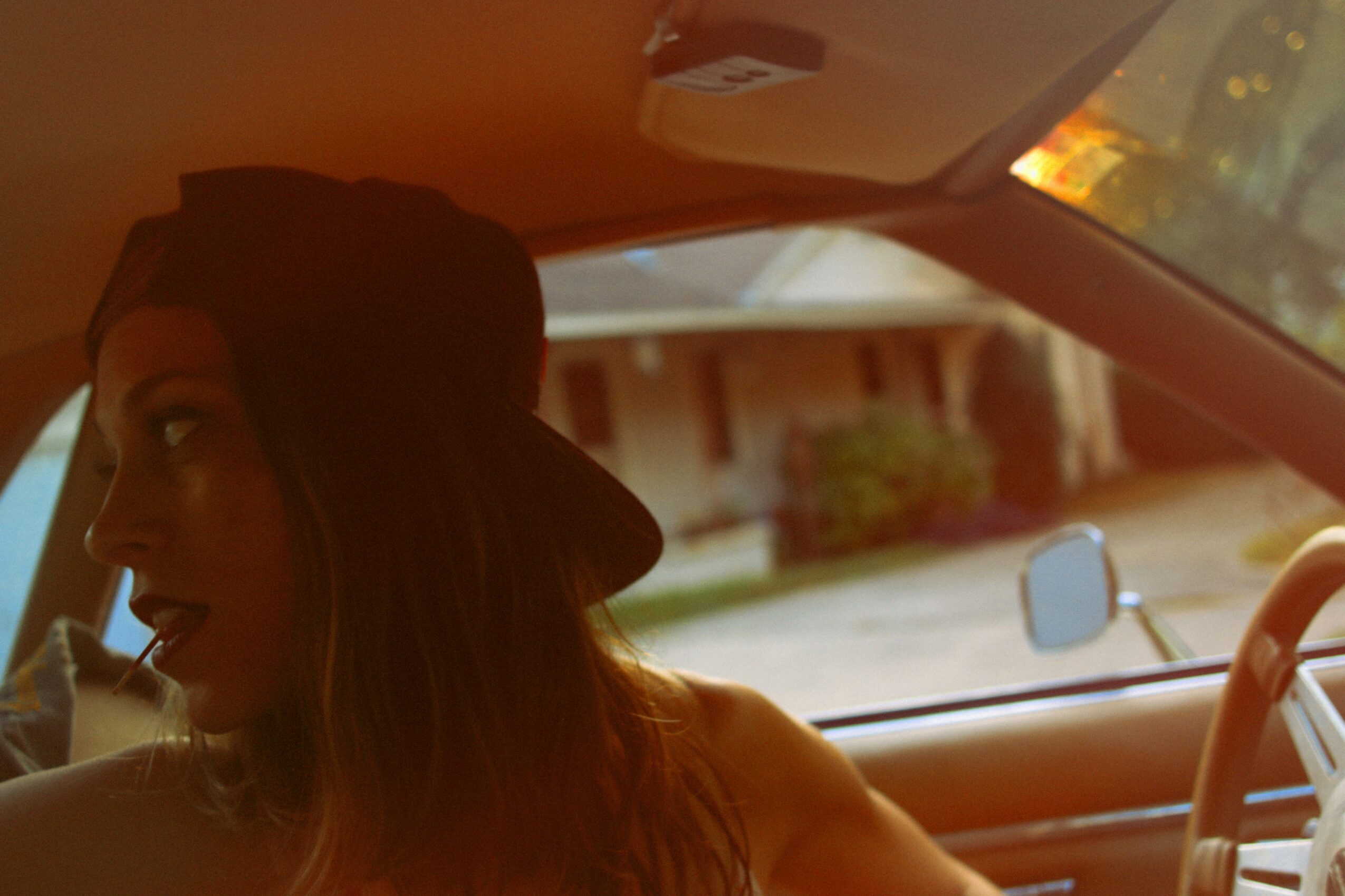 Person with long hair wearing a hat inside a car, with a residential street visible outside.