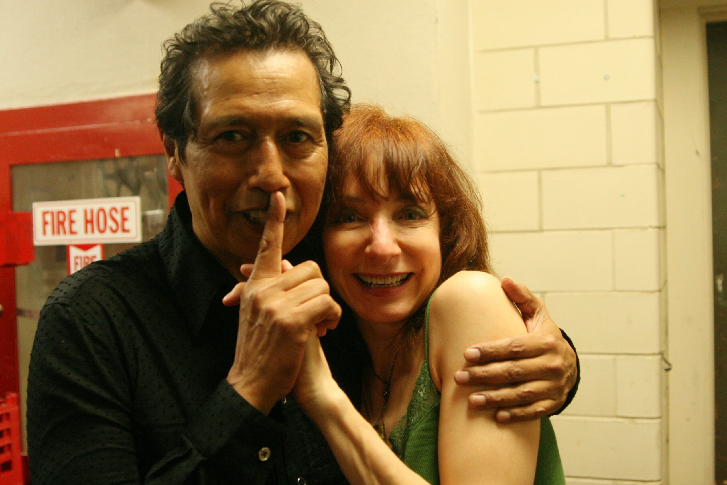 Two people smiling, one with finger on lips, hugging in front of a brick wall and fire hose sign.