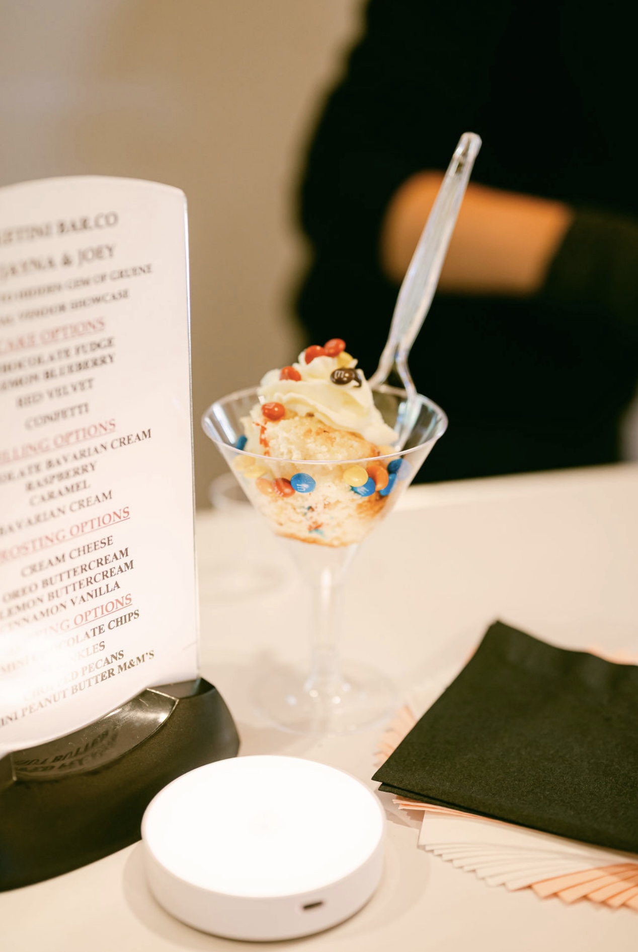 A glass dessert cup with ice cream and colorful toppings, with a spoon inside, on a table with a black napkin.