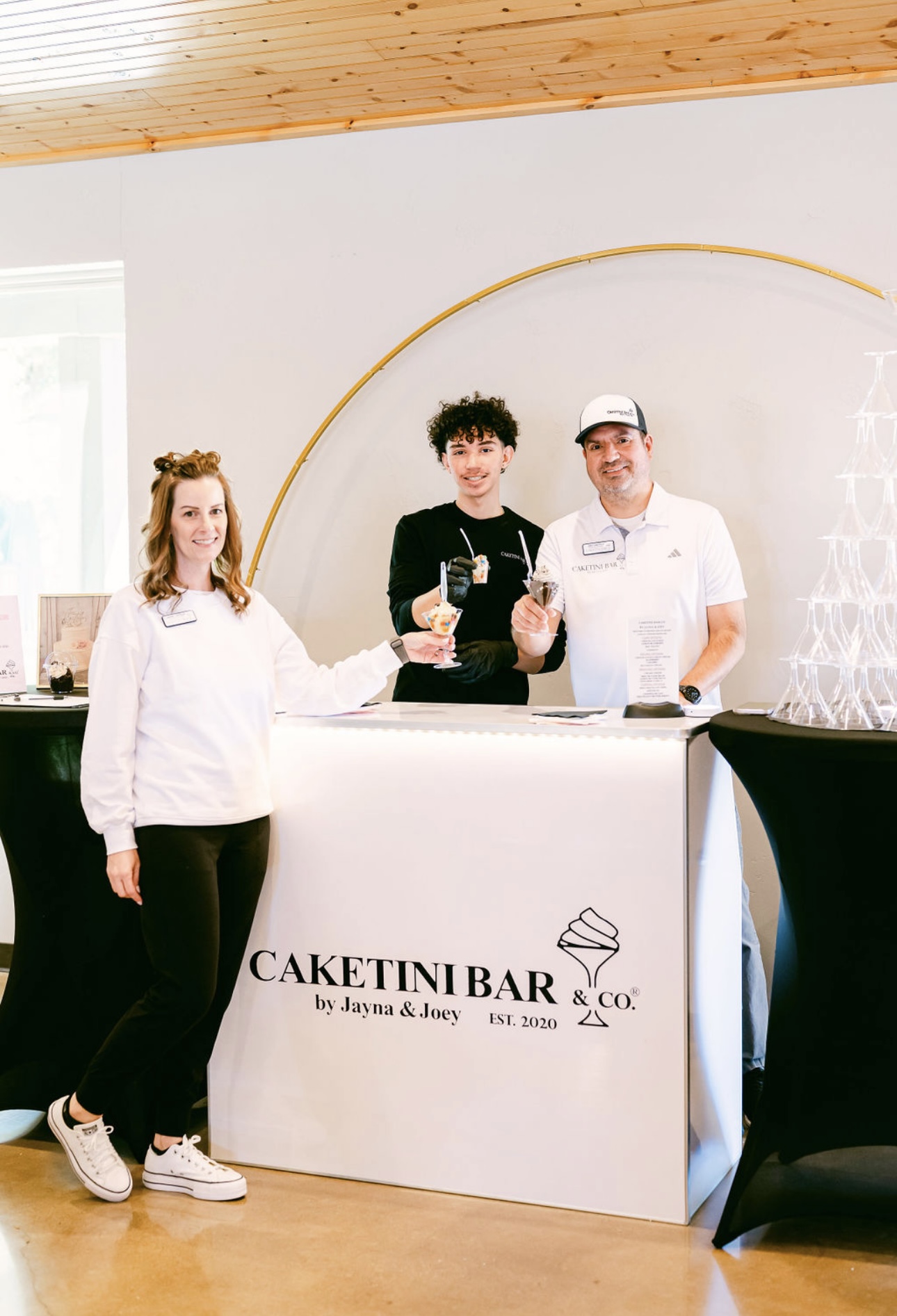 Three people behind a cake shop counter with a sign reading 'Caketini Bar & Co.' in a modern interior.