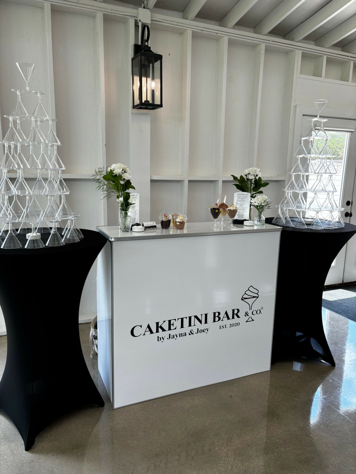 Display table with white shelves, plants, and a cake stand, with a sign reading 'Caketini Bar' in front.