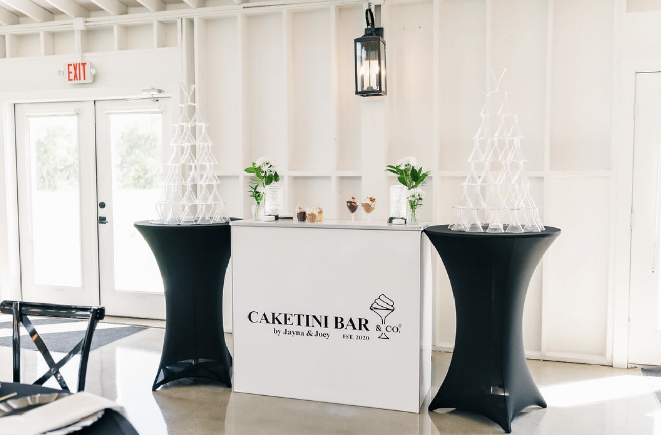 Event setup with a white table labeled 'Caketini Bar' and two black covered tables, decorated with glass and greenery, in a bright room.