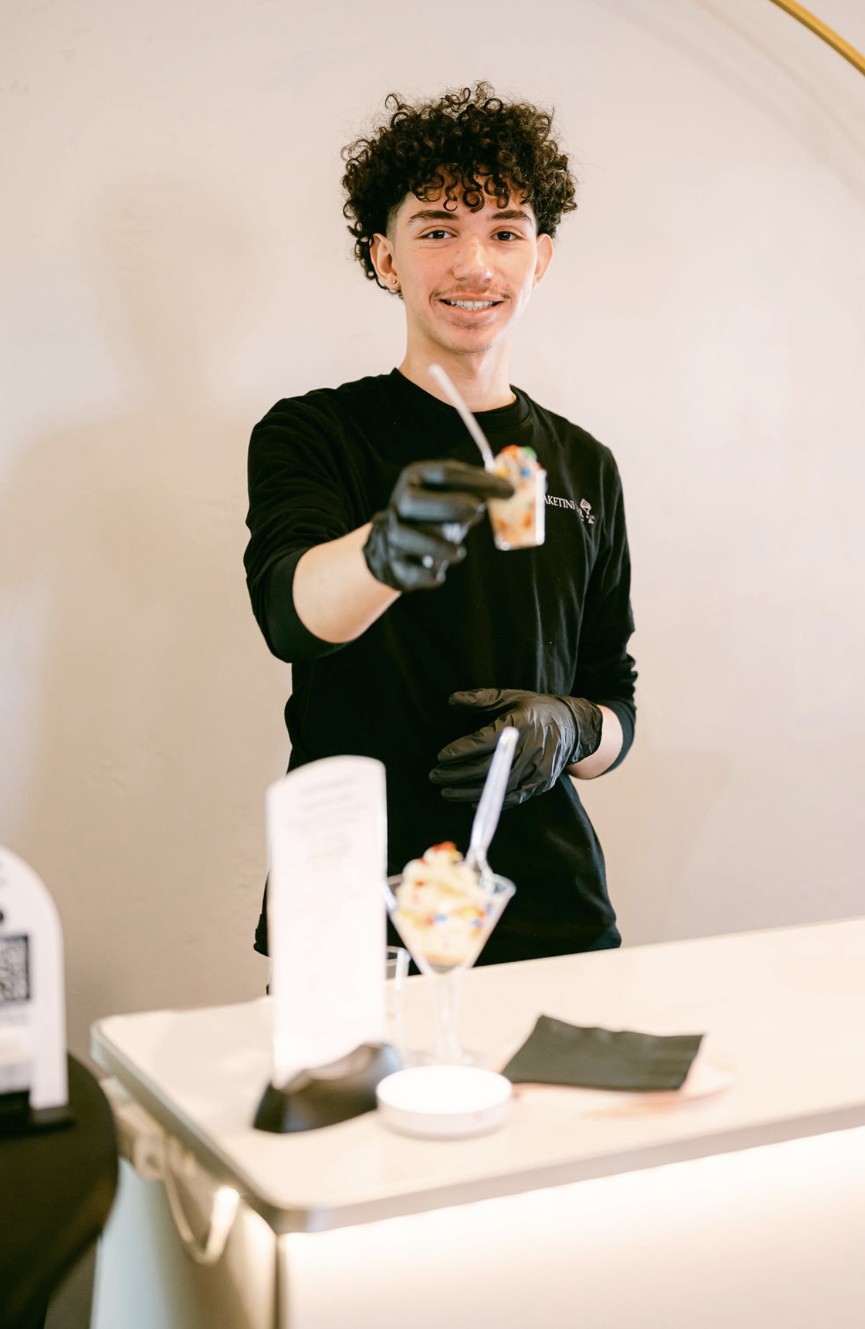 Young person with curly hair smiling, holding a dessert in a glass, wearing black gloves, standing behind a counter.