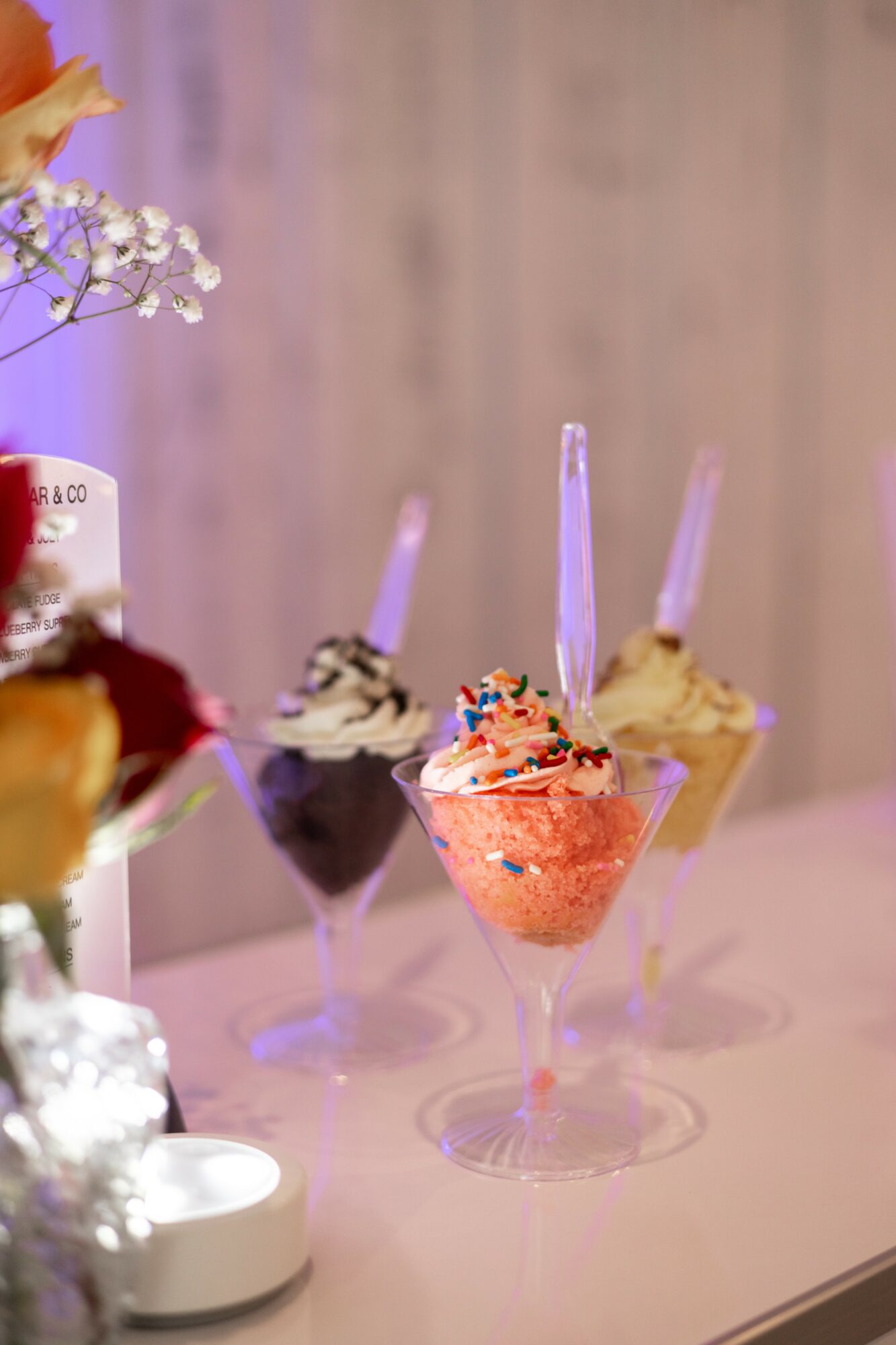 Three colorful ice cream sundaes in glass cups with sprinkles and sticks, on a white surface with a blurred background.