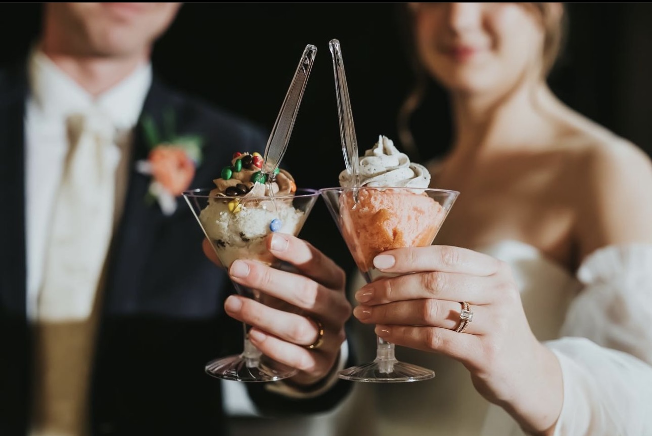 Two people holding glasses of ice cream sundaes with whipped cream and toppings, smiling in the background.