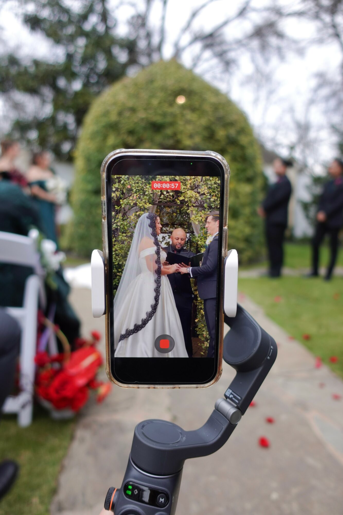 Smartphone on a stabilizer capturing a wedding ceremony outdoors with people and a large bush in the background.