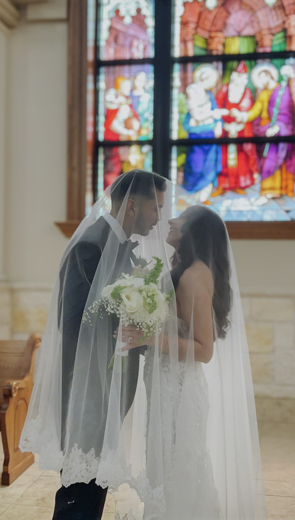 Bride and groom face each other inside a church, holding a bouquet, with a colorful stained glass window behind them.