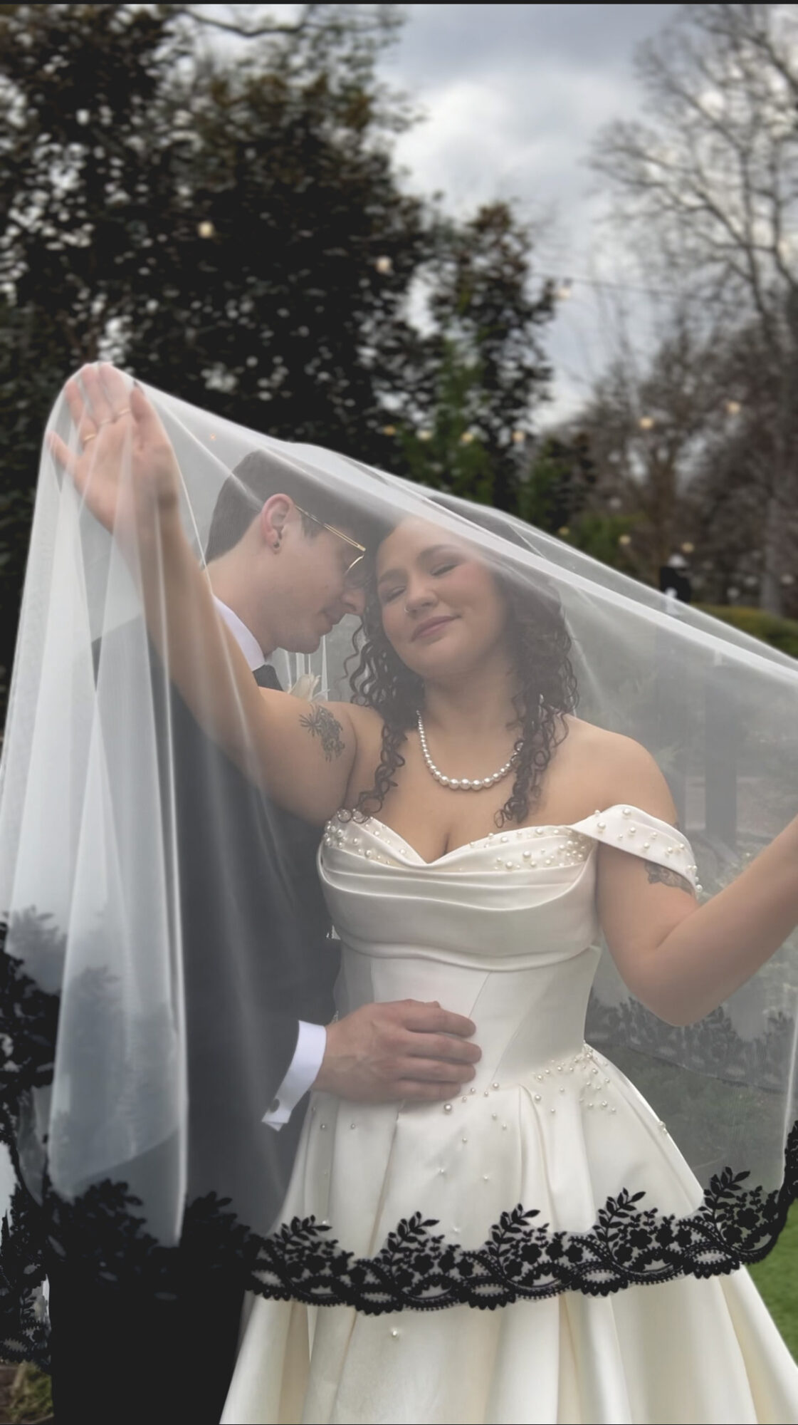 A couple in wedding attire under a transparent veil outdoors, with trees and cloudy sky in background.
