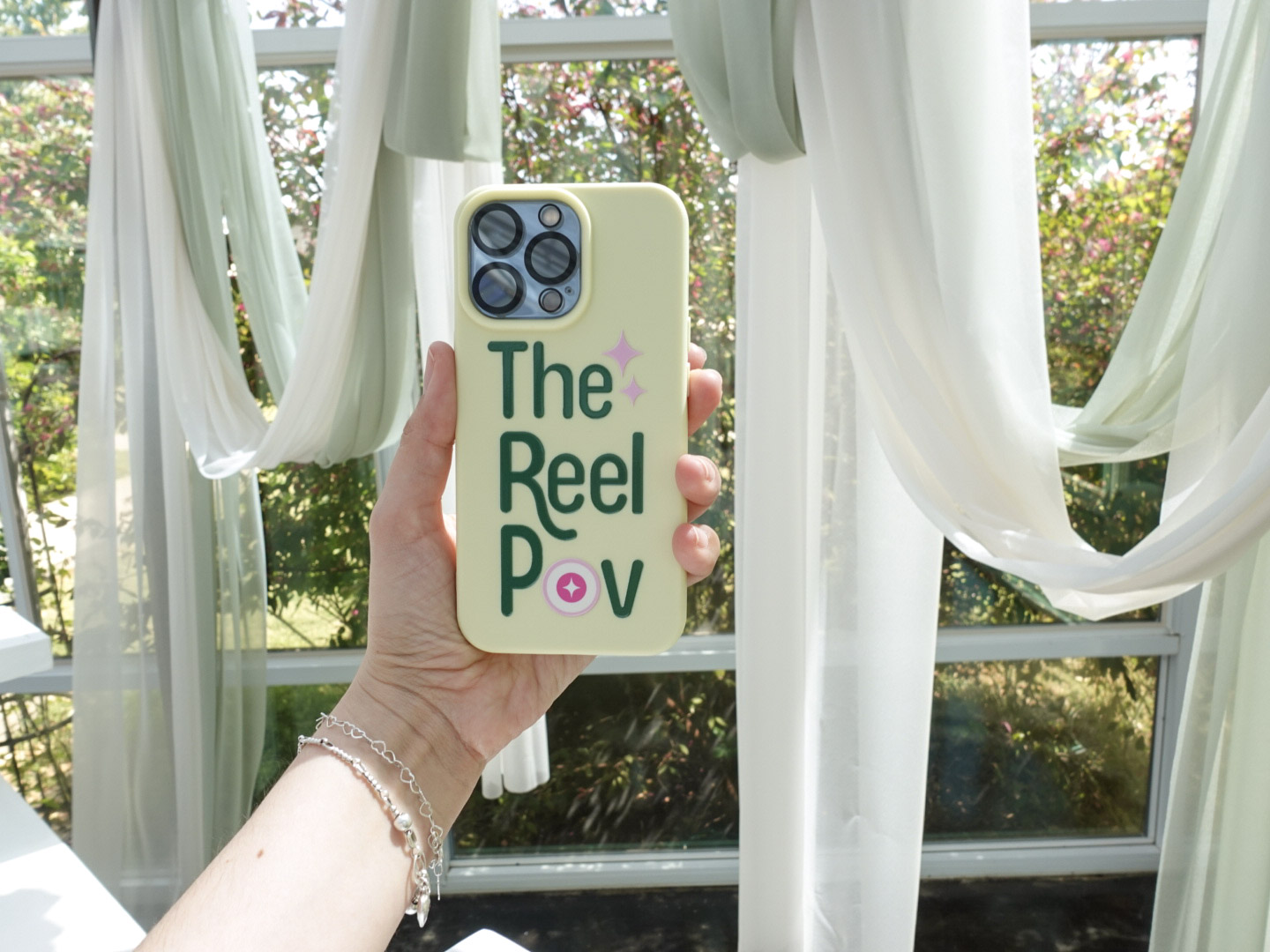 Person holding a phone with a case that says 'The Reel PoV' in front of a window with white curtains and greenery outside.