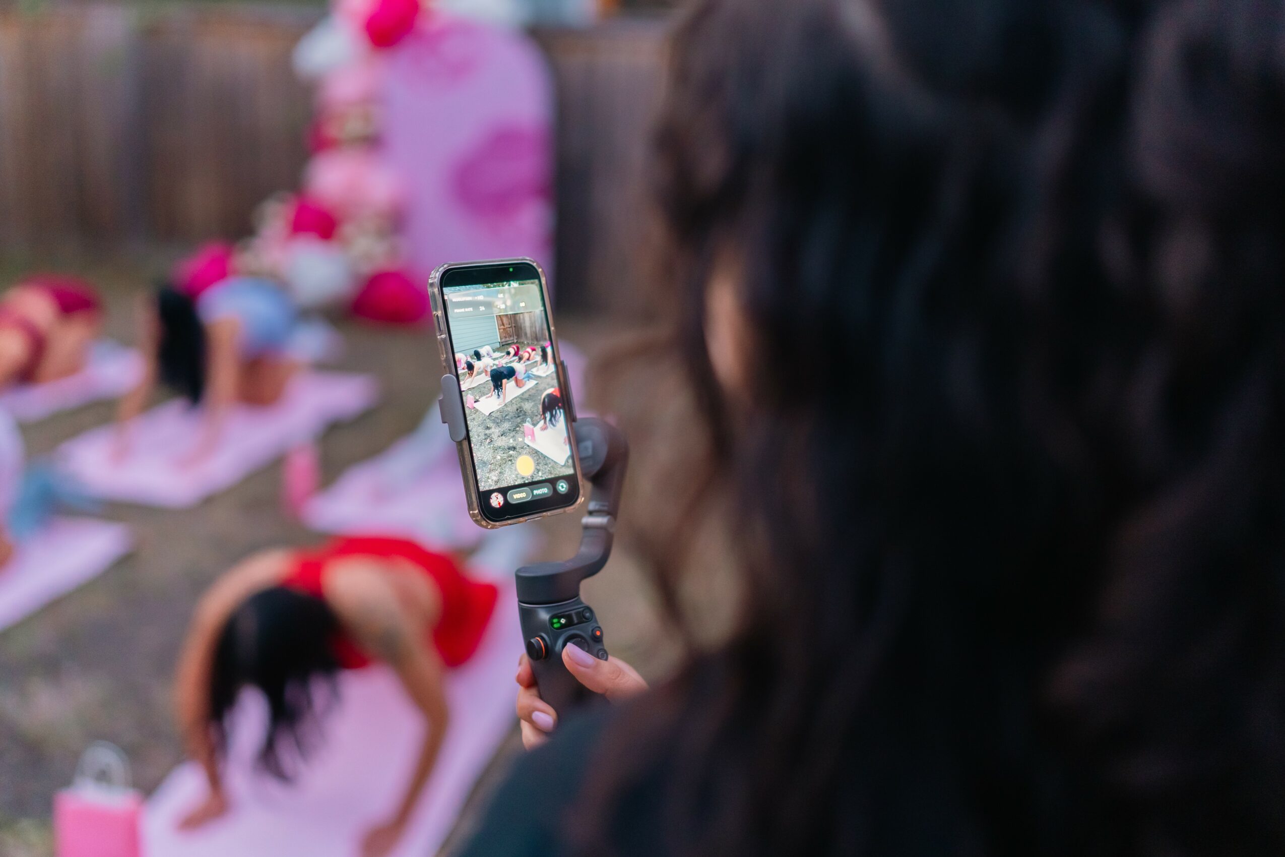 Person taking a photo with a smartphone of children playing outdoors, with a pink decoration in the background.