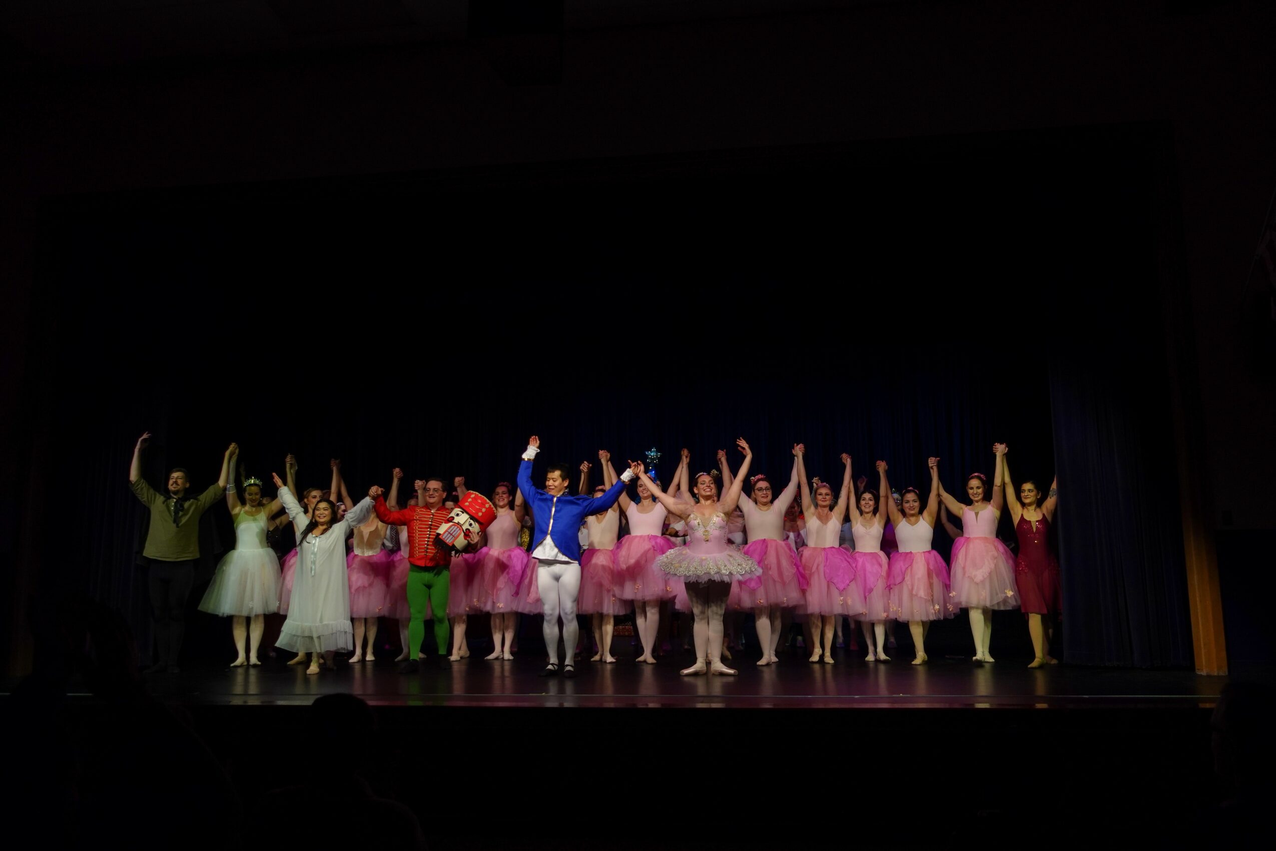 Group of ballet dancers on stage, some in pink tutus, others in colorful costumes, raising arms in performance.