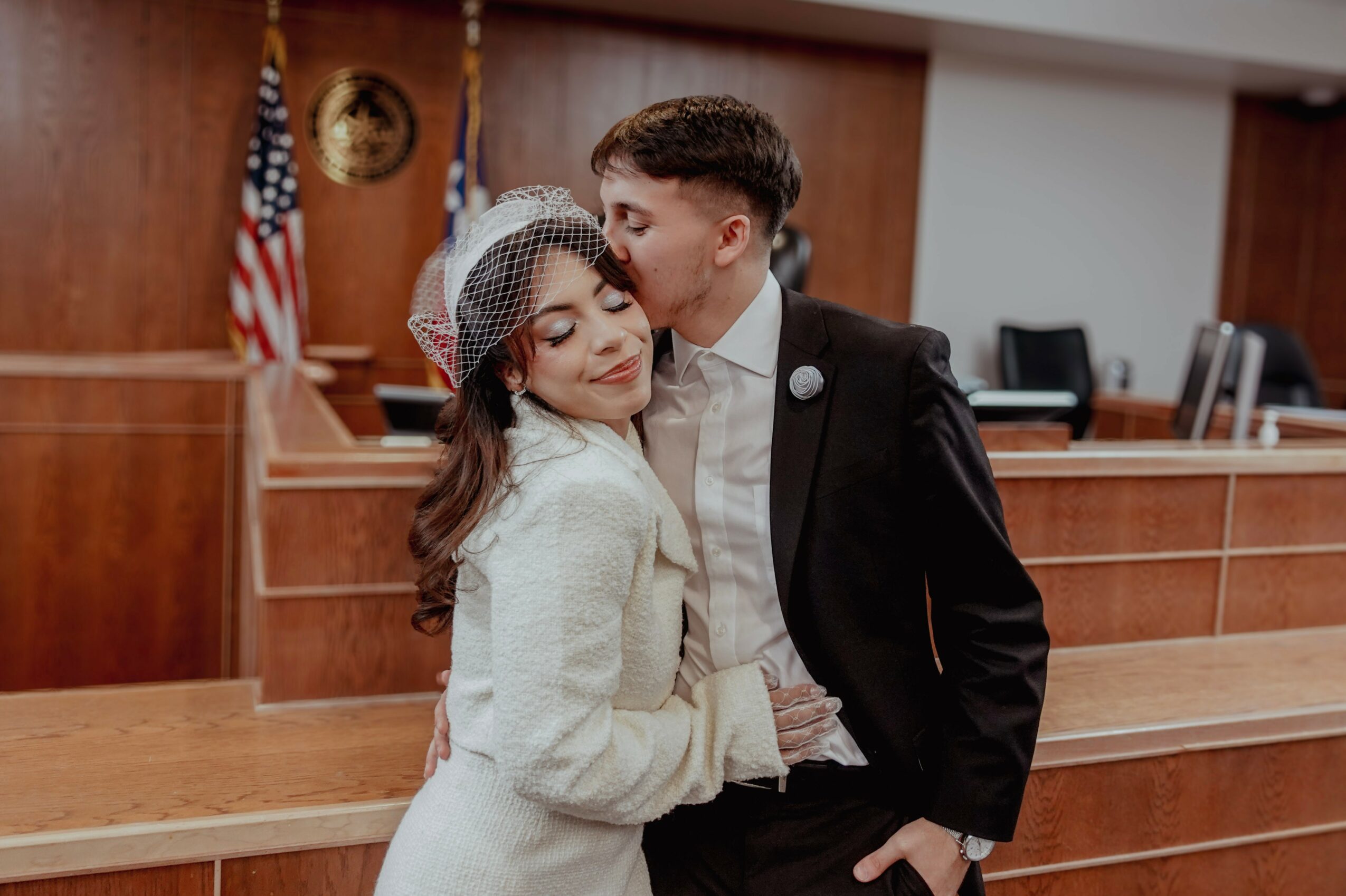 A man kisses a woman on the cheek in a courtroom, both dressed formally, with American flags in the background.