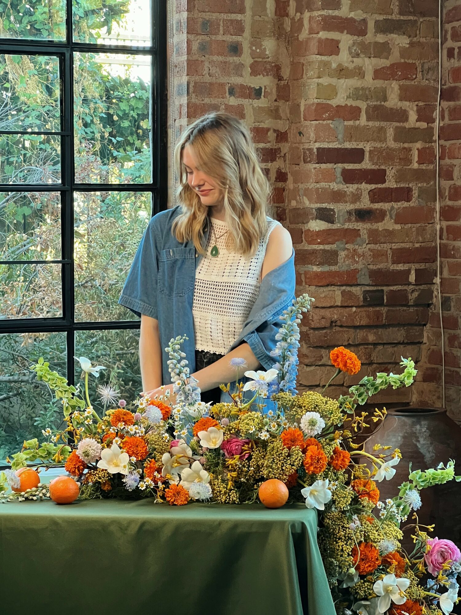 Young woman with blonde hair standing behind a table with colorful flowers and oranges, in a room with brick wall and large window.