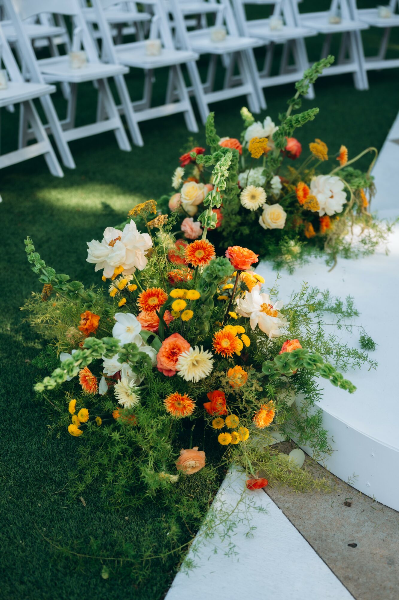 Colorful flower arrangements with white, orange, and yellow flowers on a white platform outdoors, with white chairs in the background.