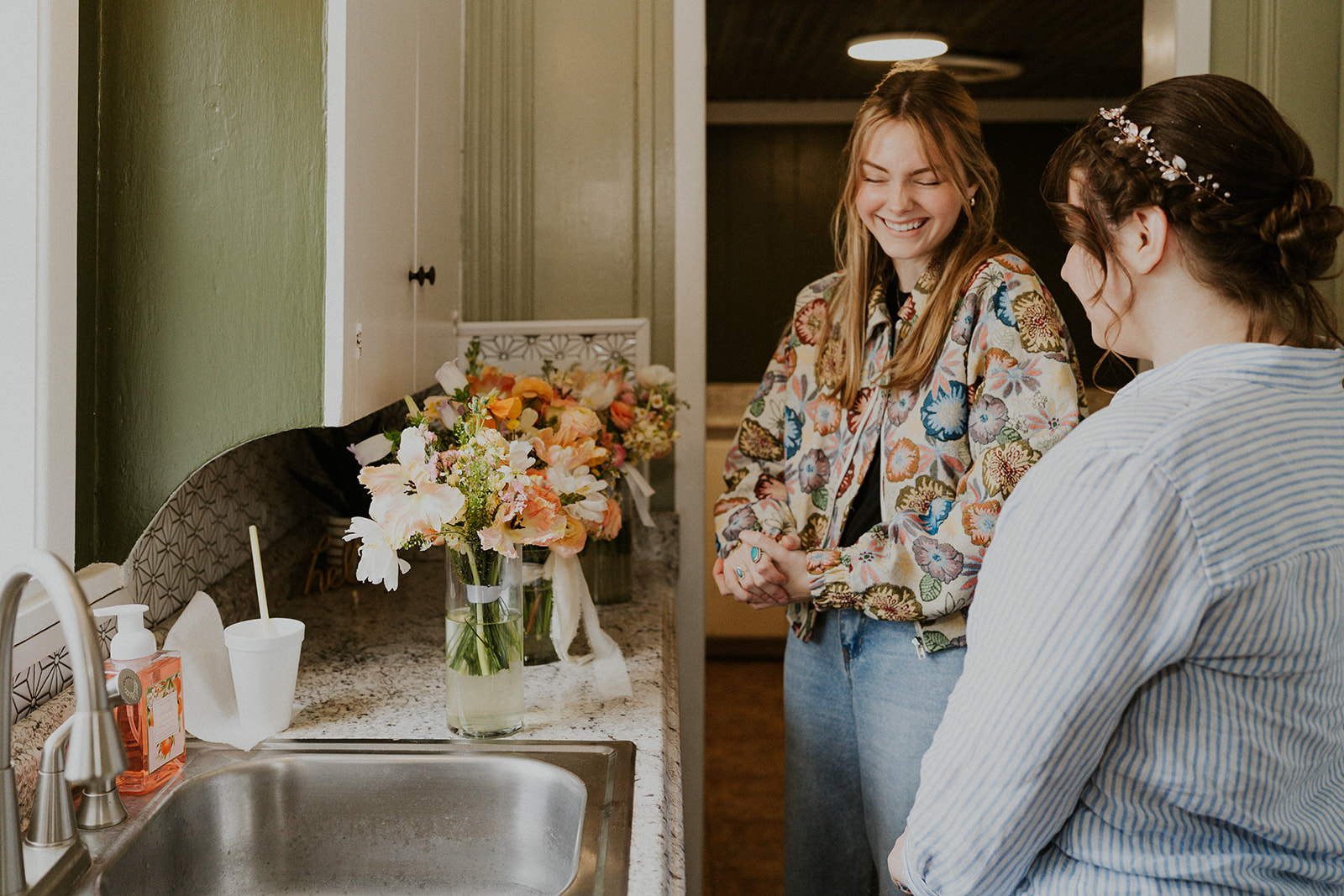 Two women smiling and talking in a kitchen with flowers on the counter, one wearing a patterned shirt and the other in a striped sweater.