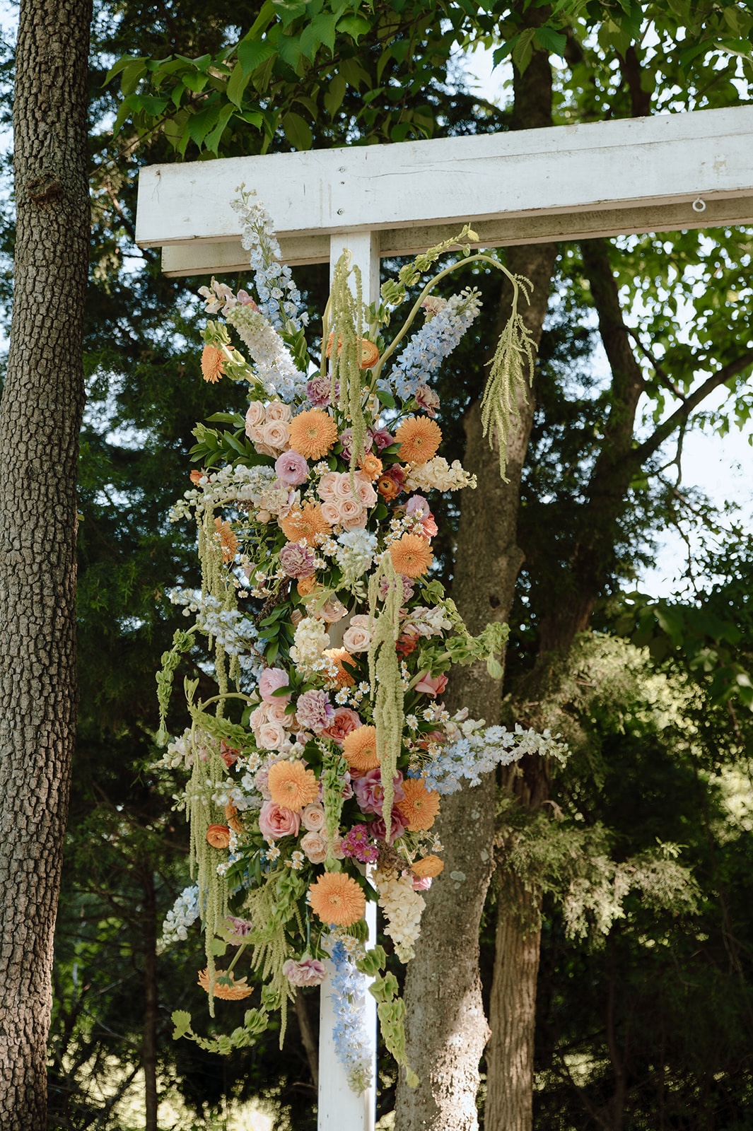 Floral arrangement with pink, peach, and white flowers hanging from a white structure outdoors, surrounded by trees and greenery.