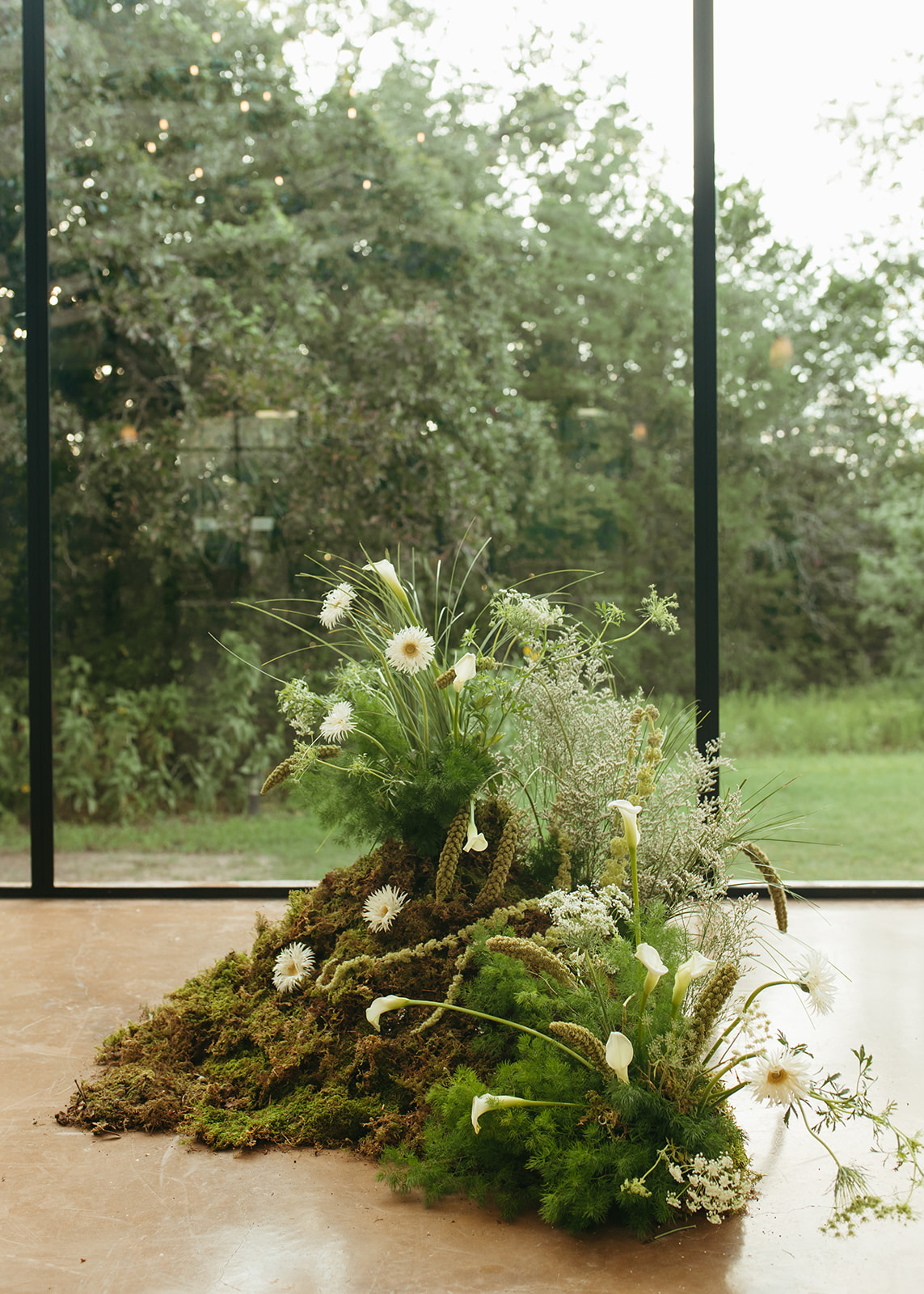 Indoor floral arrangement with white flowers and greenery on moss, in front of large window showing trees outside.