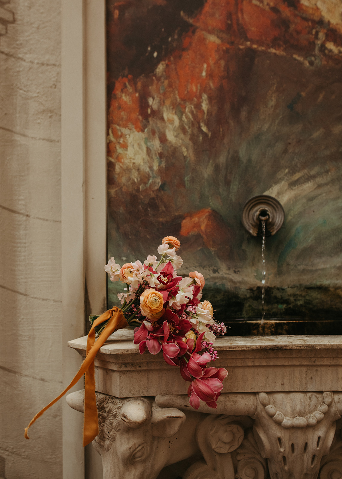 A floral arrangement with pink, white, and orange flowers and a ribbon, placed on a decorative stone fountain with a water spout, against a painted wall.