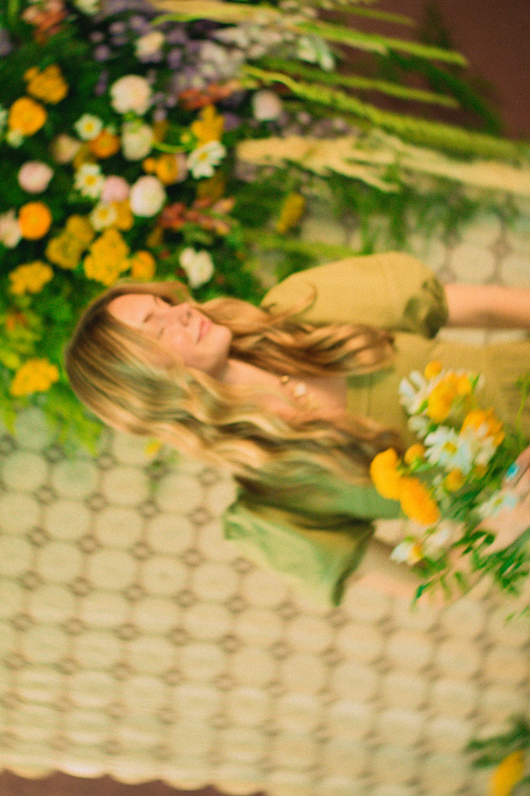 Young woman with long hair holding a bouquet of yellow and white flowers, standing near a floral arrangement, smiling.