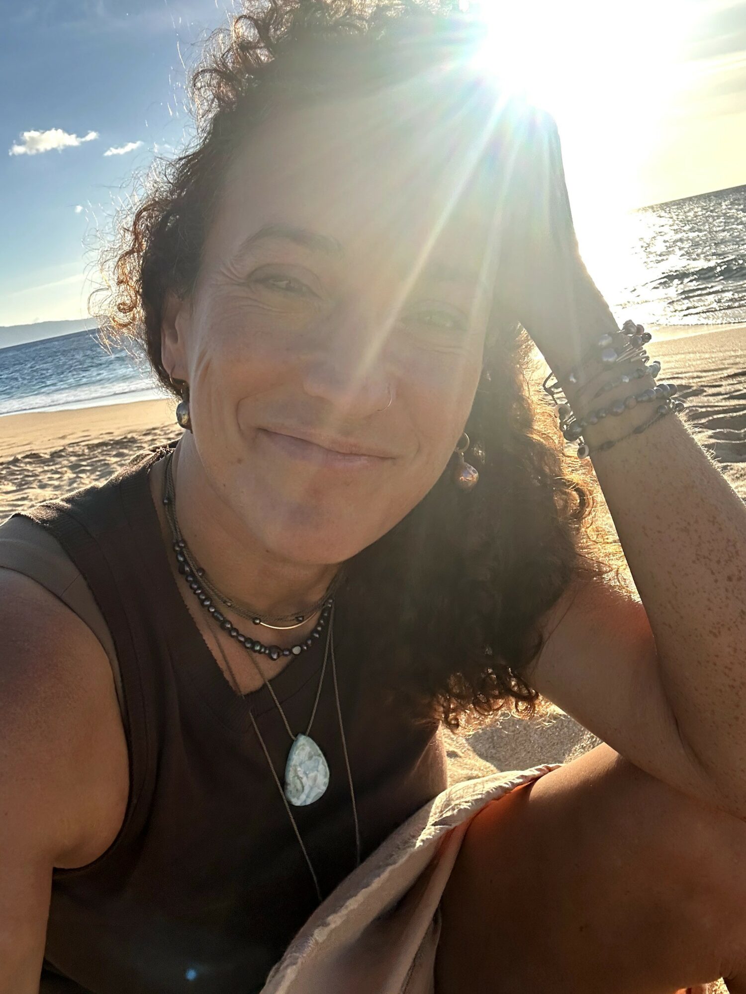 Woman smiling at the beach with sunlight shining behind her, ocean and sky in the background.