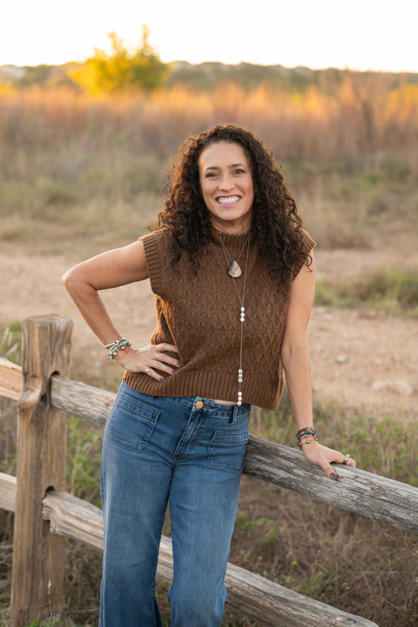 Woman with curly hair smiling outdoors, leaning on a wooden fence, wearing a brown sleeveless top and blue jeans.