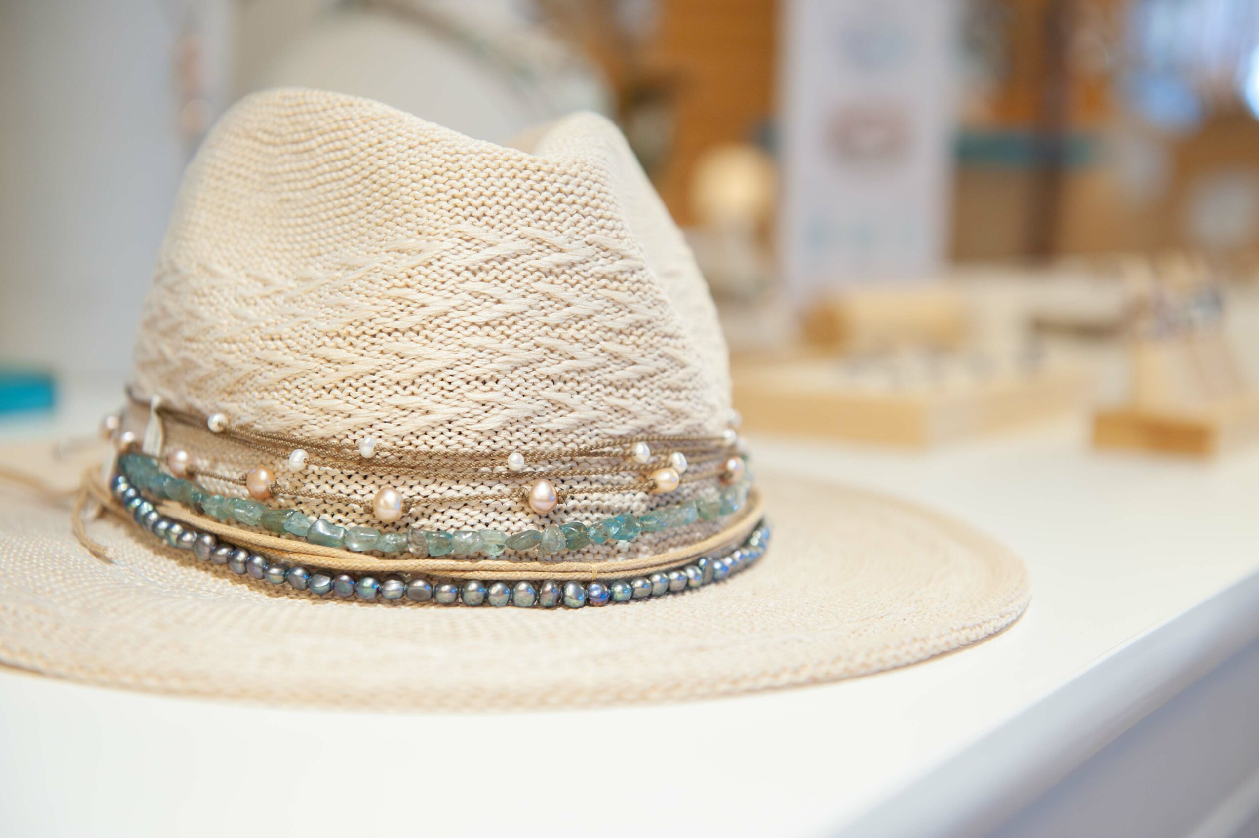 A beige woven hat with a decorative band of beads and pearls, placed on a white surface in a store setting.