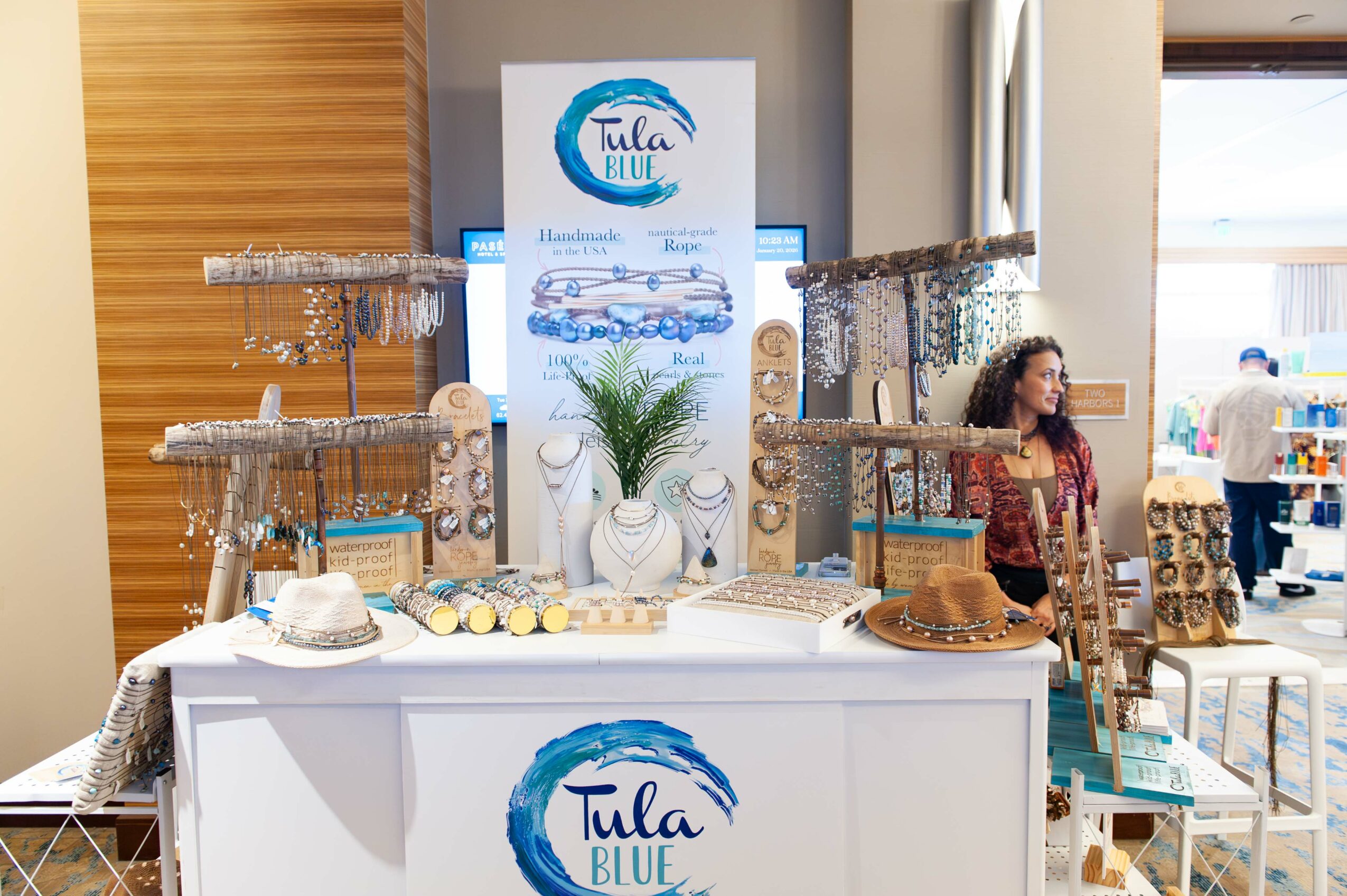 Display table with Tula Blue logo, decorated with a plant, cakes, and glasses, in a bright room with people and windows.
