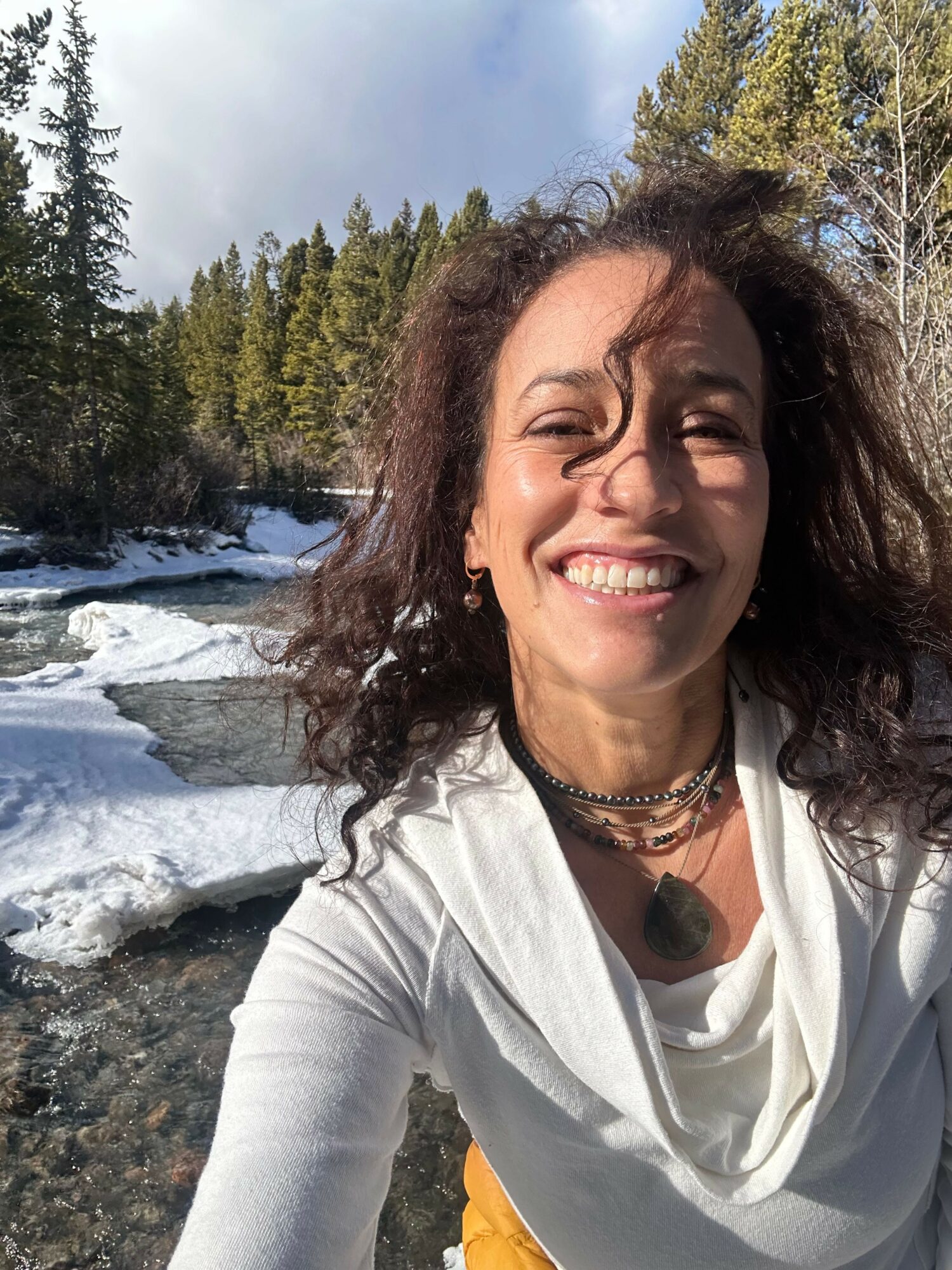 Woman with curly hair smiling outdoors near a river with trees and rocks in the background.