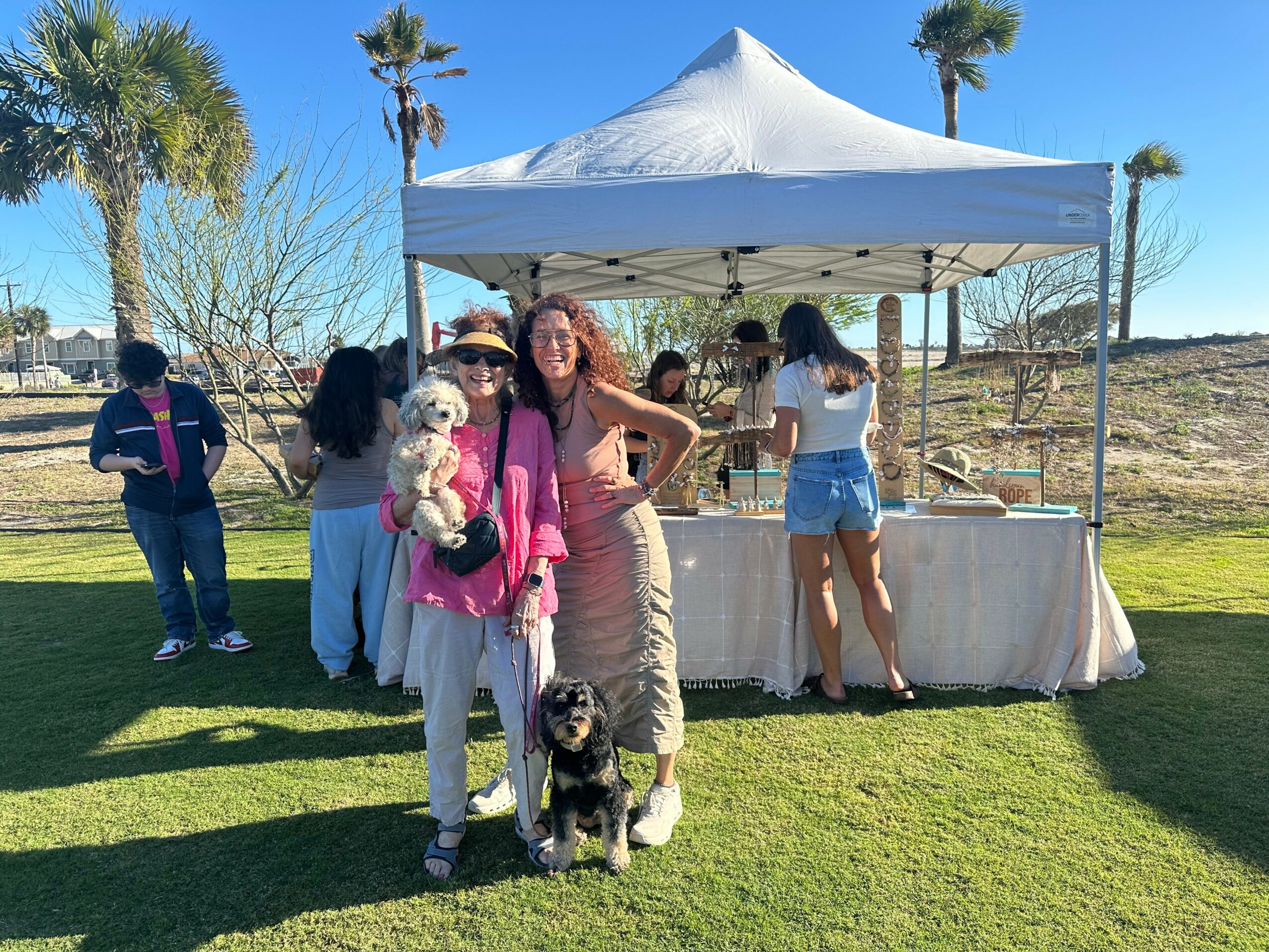 Two women and two dogs in front of a white canopy tent outdoors on grass, with trees and a clear blue sky.