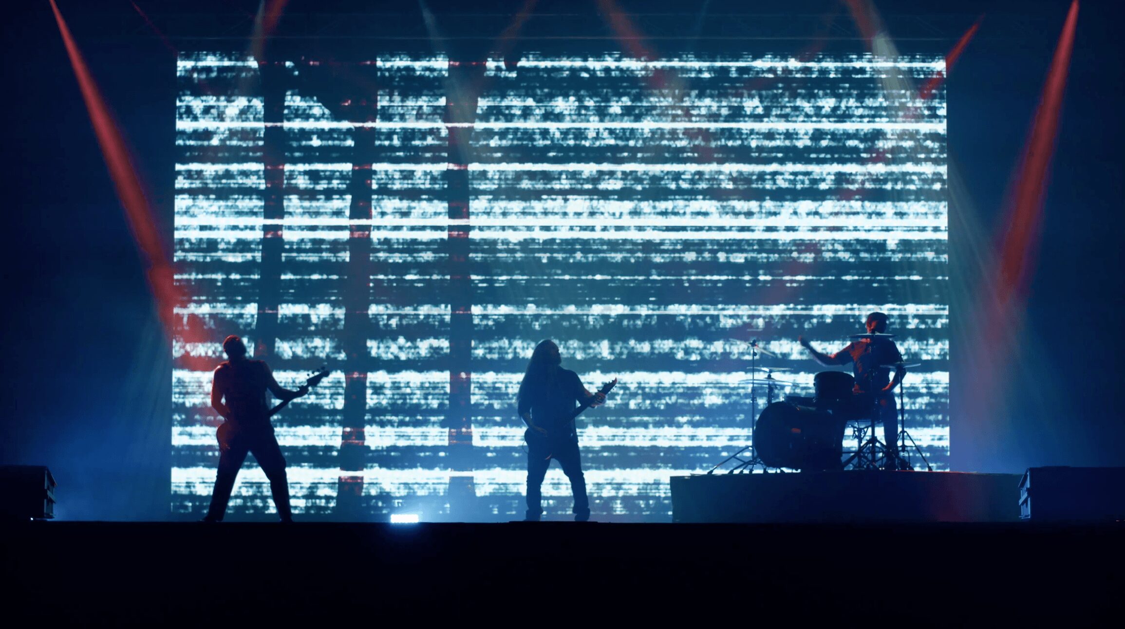 Three musicians perform on stage with a large digital screen displaying horizontal lines behind them.